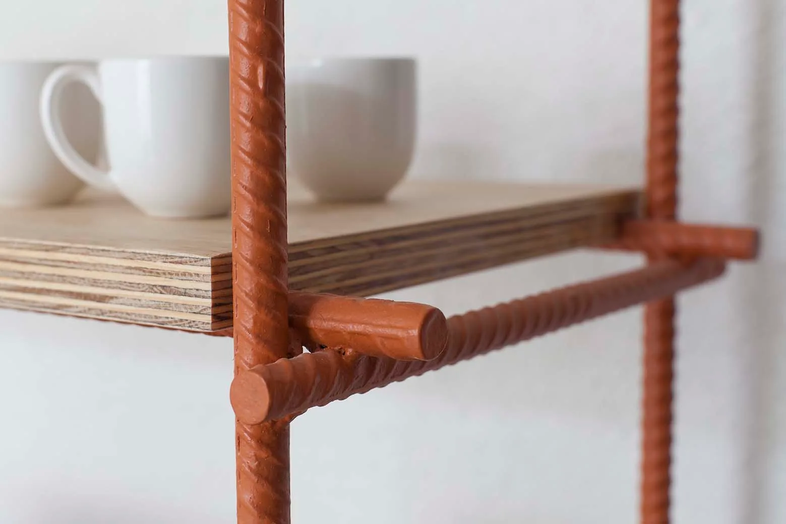 A minimalist shelving unit features a plywood shelf supported by a terracotta orange painted rebar frame against a white wall.