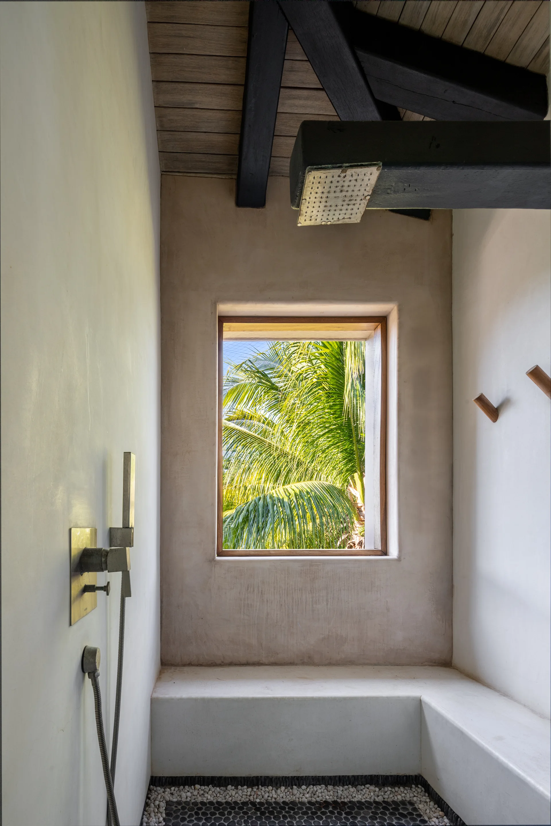 A walk-in plaster shower features a square window framing palm trees, a built-in stucco bench, dark wooden ceiling beams, and a pebbled floor.