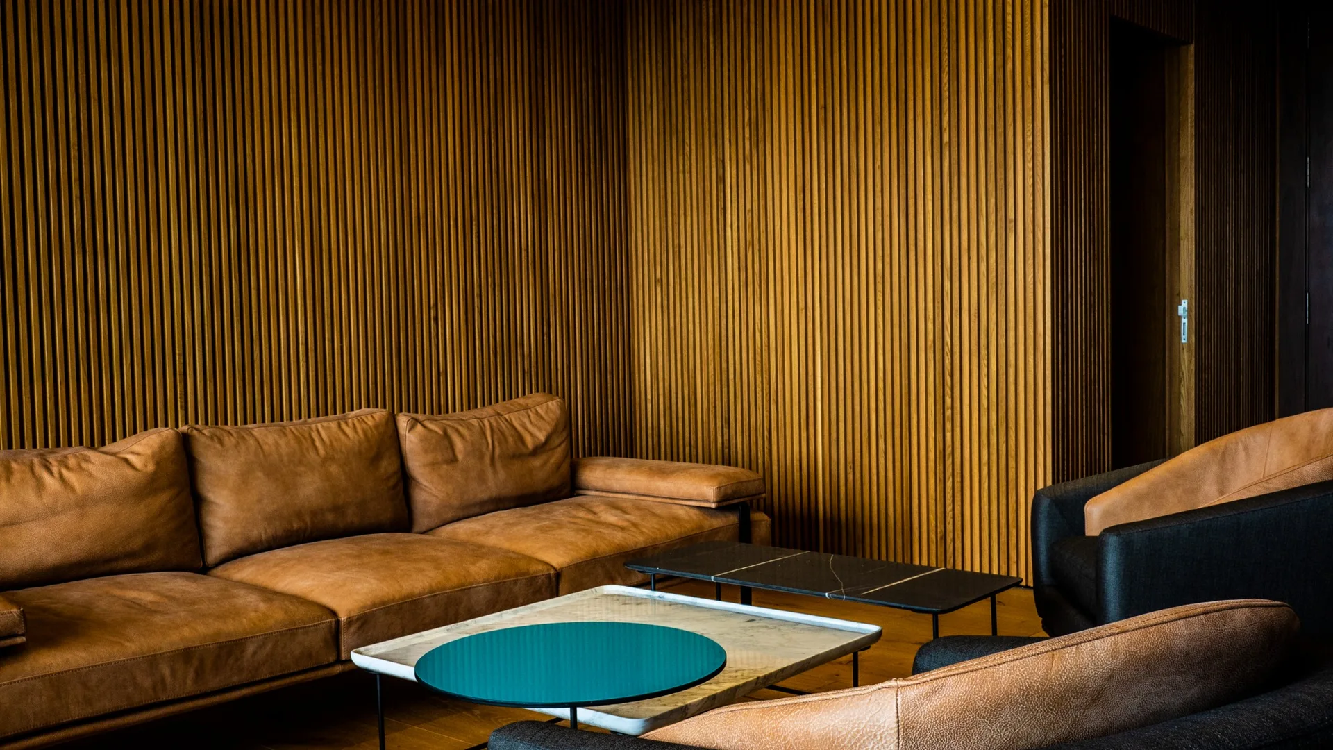 A modern living room features a tan leather sofa and armchairs arranged against a wall covered in vertical slatted wood paneling.