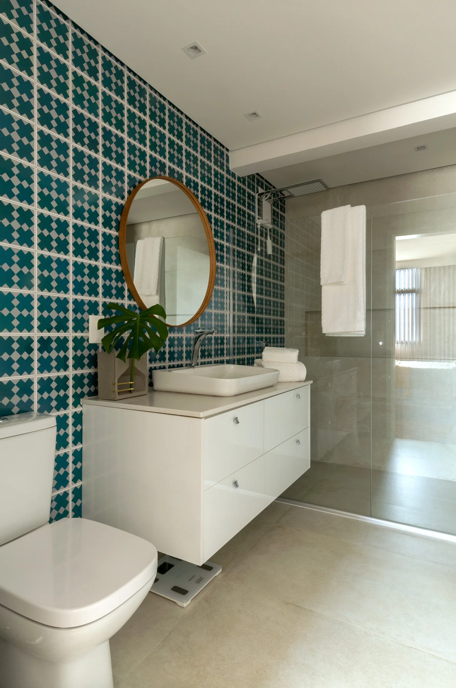 A modern bathroom features a teal and white geometric tile wall behind a white floating vanity, a round wooden mirror, and a glass-enclosed shower.