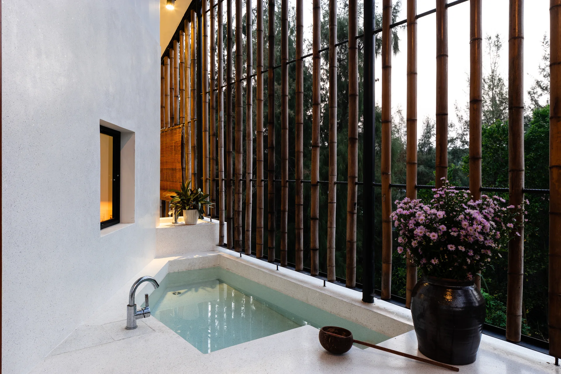 A sunken soaking tub is surrounded by white terrazzo stone alongside a wall of vertical bamboo poles filtering natural light.