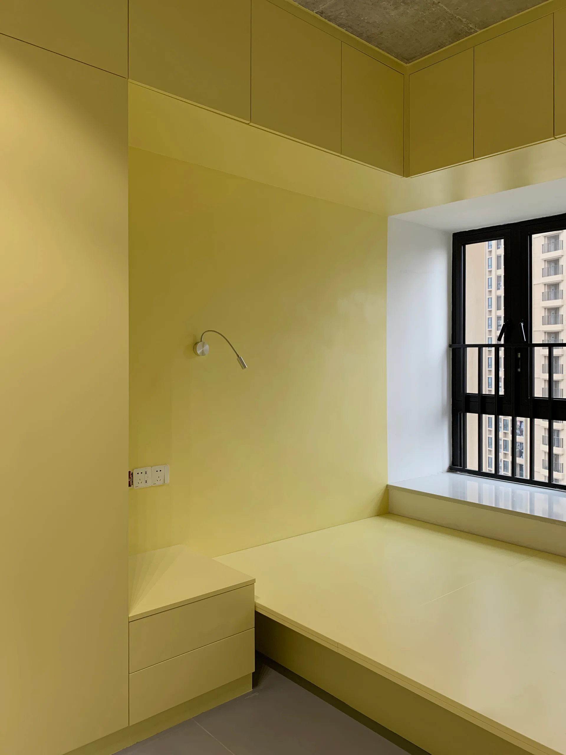 A minimalist bedroom features a custom yellow built-in platform bed and floor-to-ceiling storage cabinets next to a black-framed window.