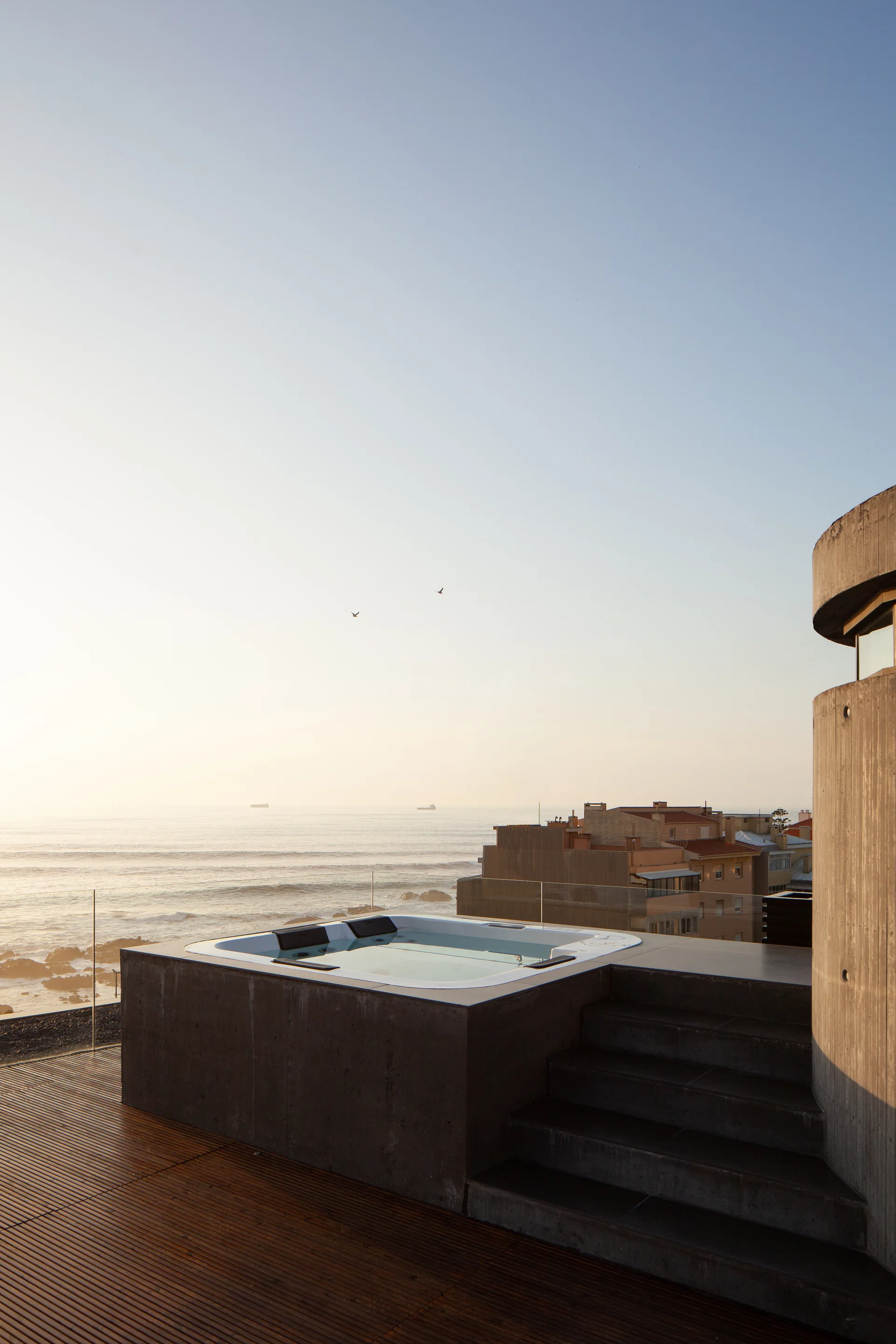 A modern outdoor hot tub built into a concrete block sits on a wooden deck next to a glass railing overlooking the ocean.