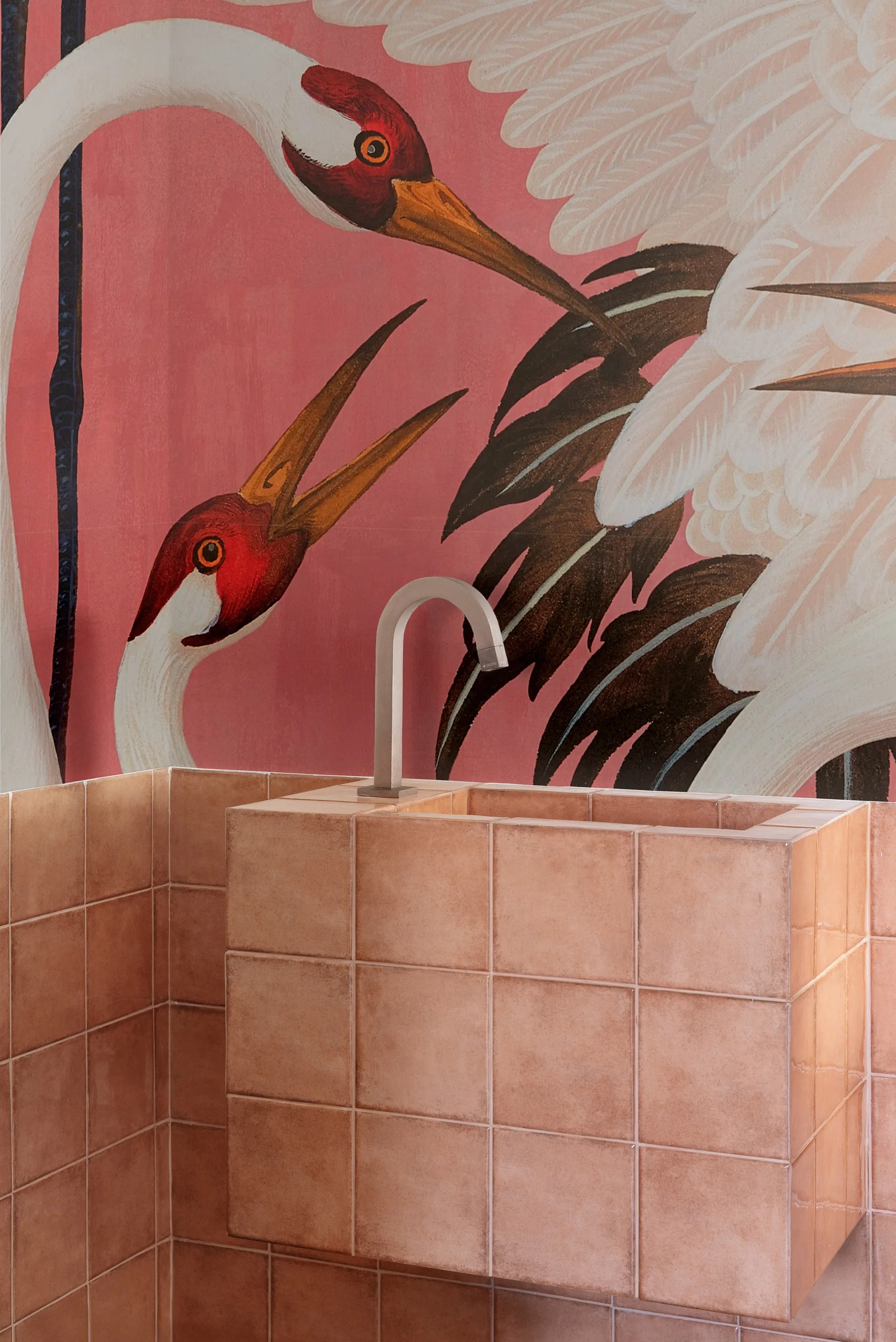 A bathroom featuring a square basin covered in salmon-colored ceramic tiles beneath a brushed metal faucet, set against a pink crane wall mural.