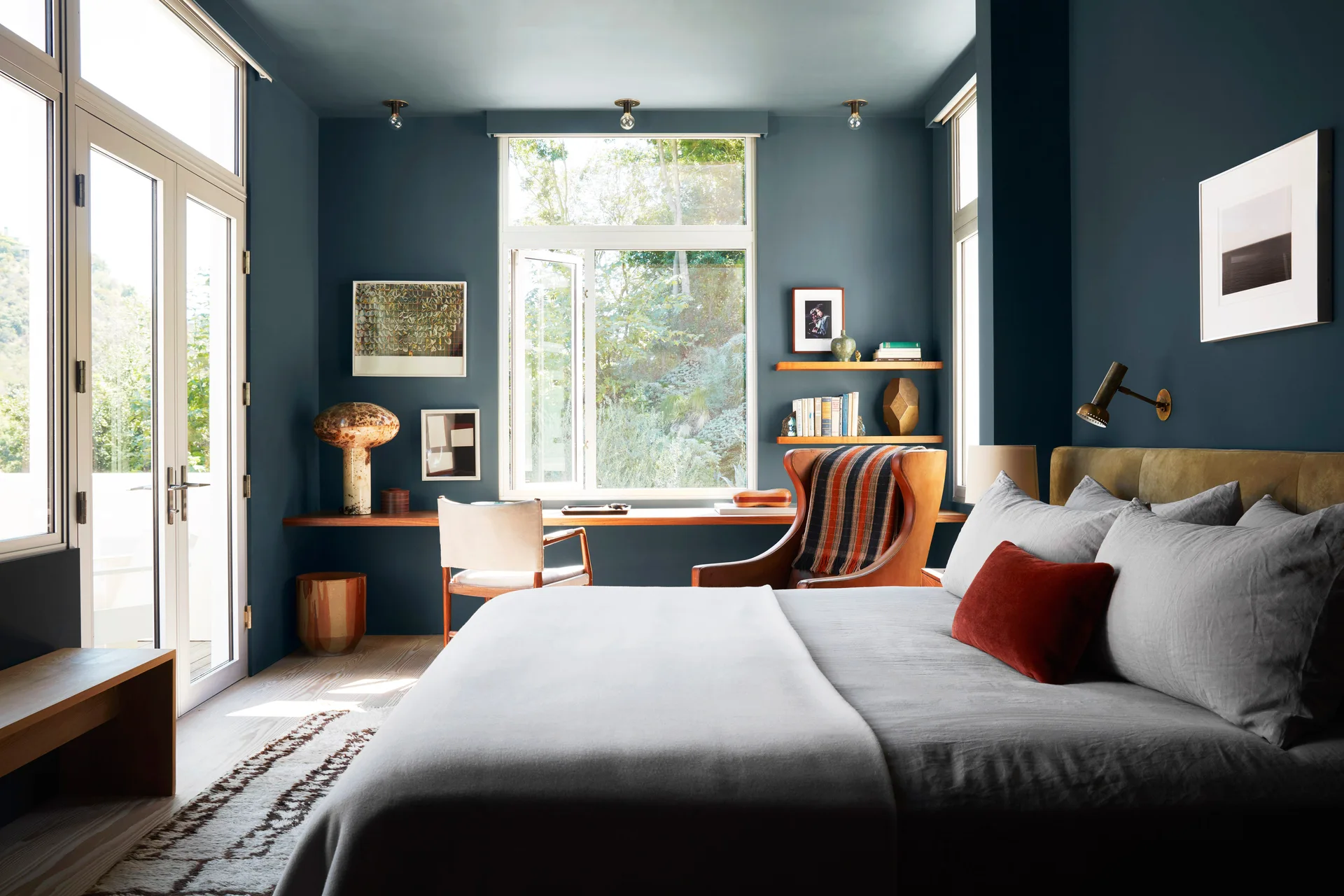 A moody bedroom features dark blue walls and ceiling, a built-in wooden desk under a large window, and an upholstered bed with gray linens.