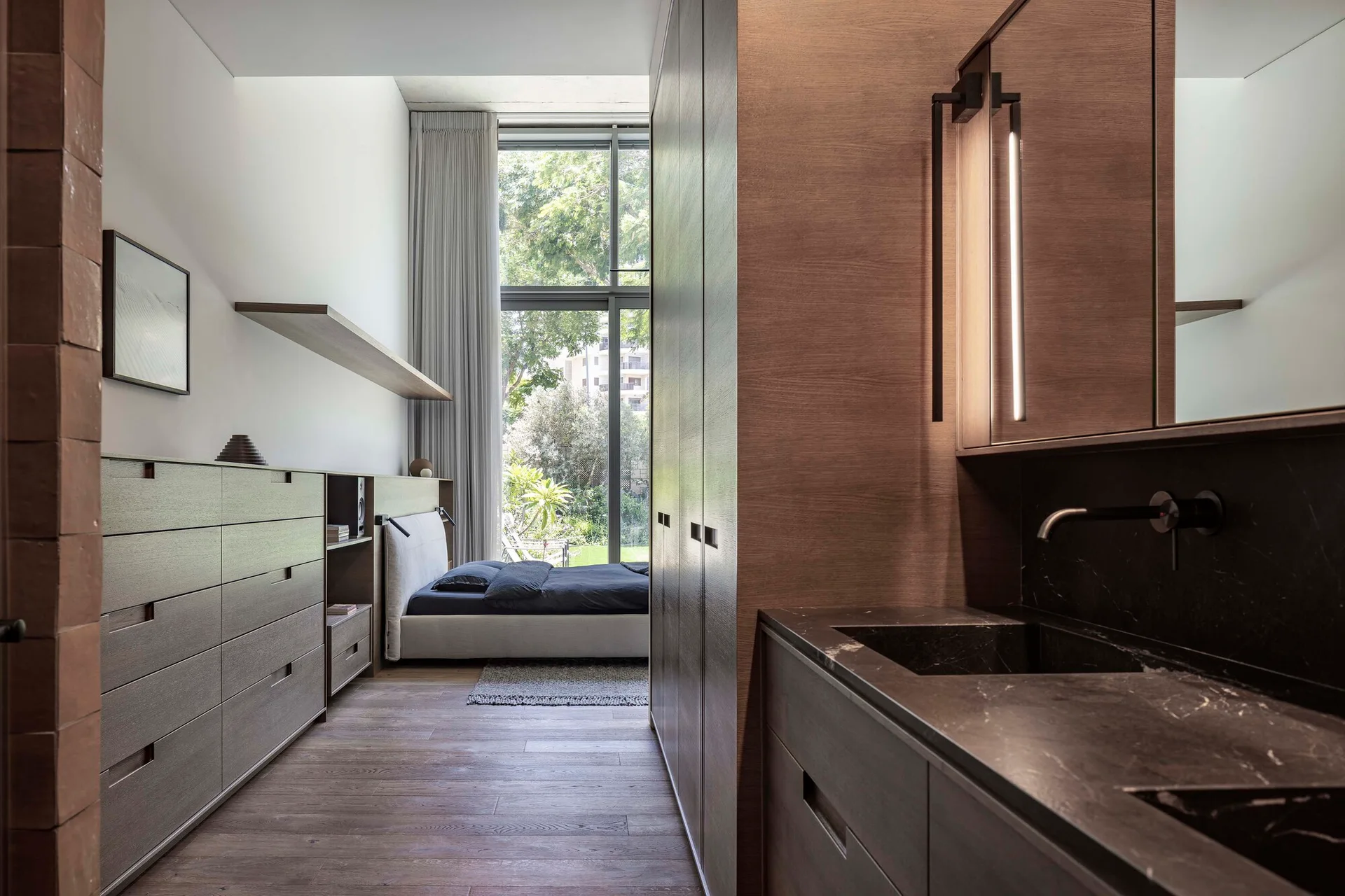 An integrated wooden wardrobe and dark stone vanity line a hallway that opens into a minimalist bedroom with a large garden-facing window.