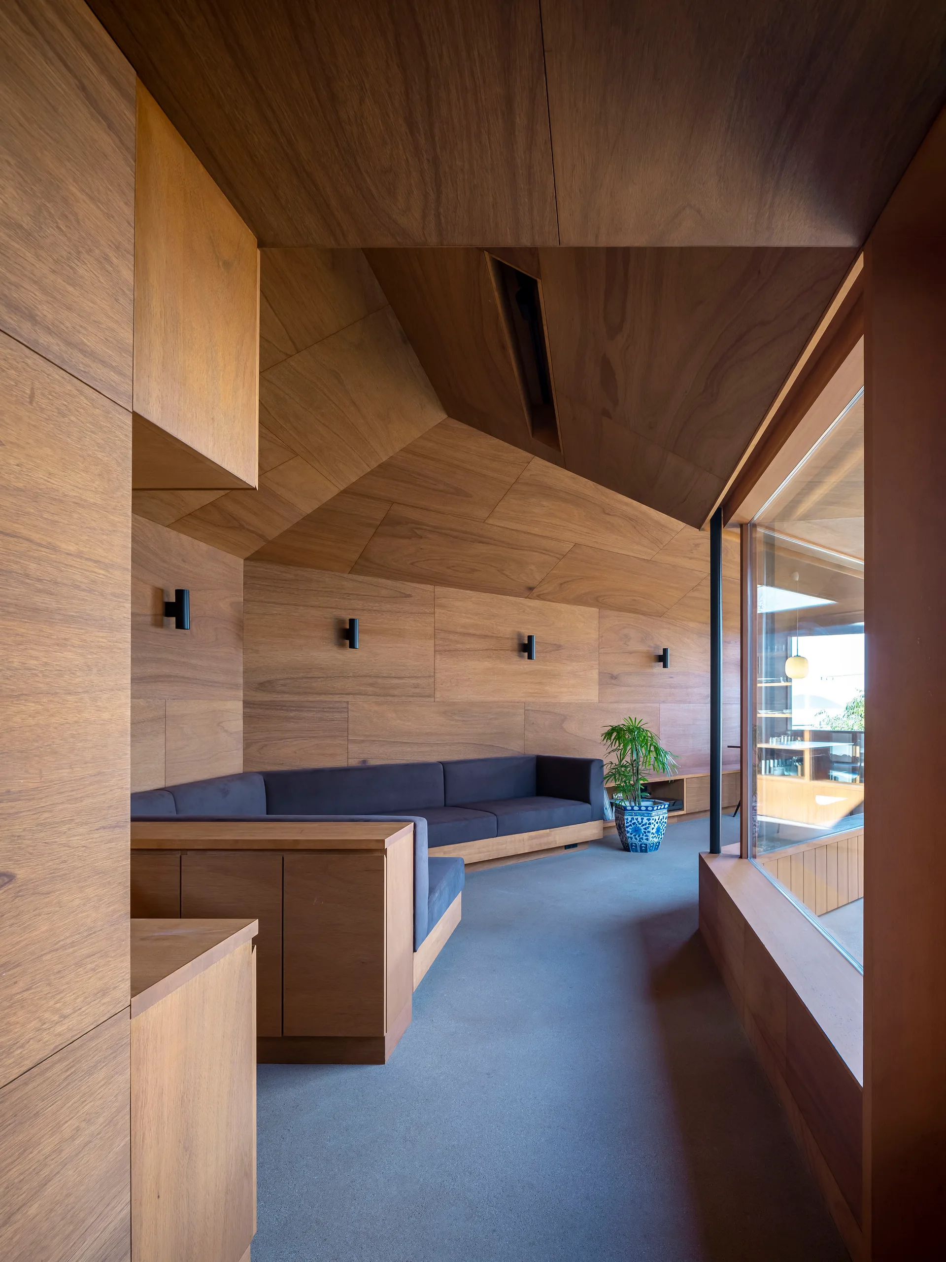 A minimalist room with warm wood paneled walls and ceiling, featuring a custom built-in gray corner sofa and a concrete floor.