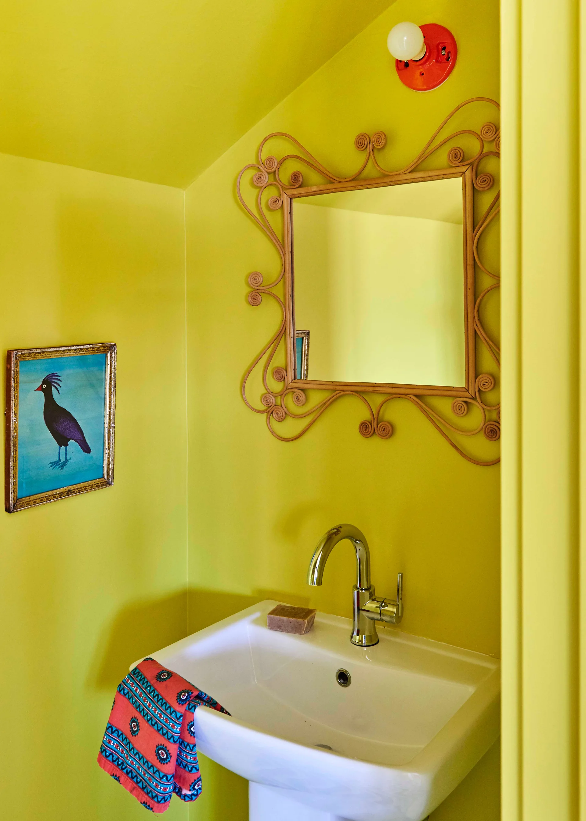 A bright yellow powder room features a white pedestal sink, a rattan-framed wall mirror, and a small framed illustration of a bird.