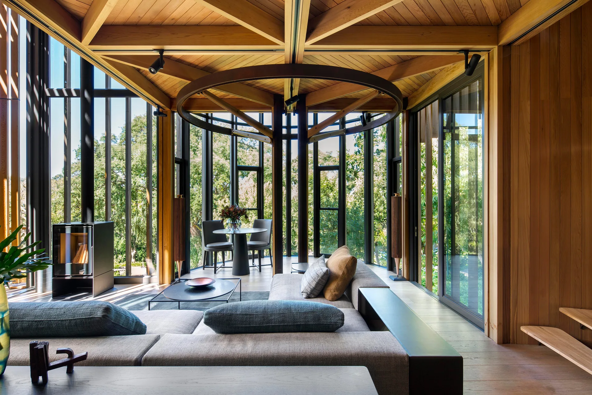 A modern living room features a modular grey sofa, a freestanding black fireplace, and a geometric wood-paneled ceiling surrounded by glass walls.