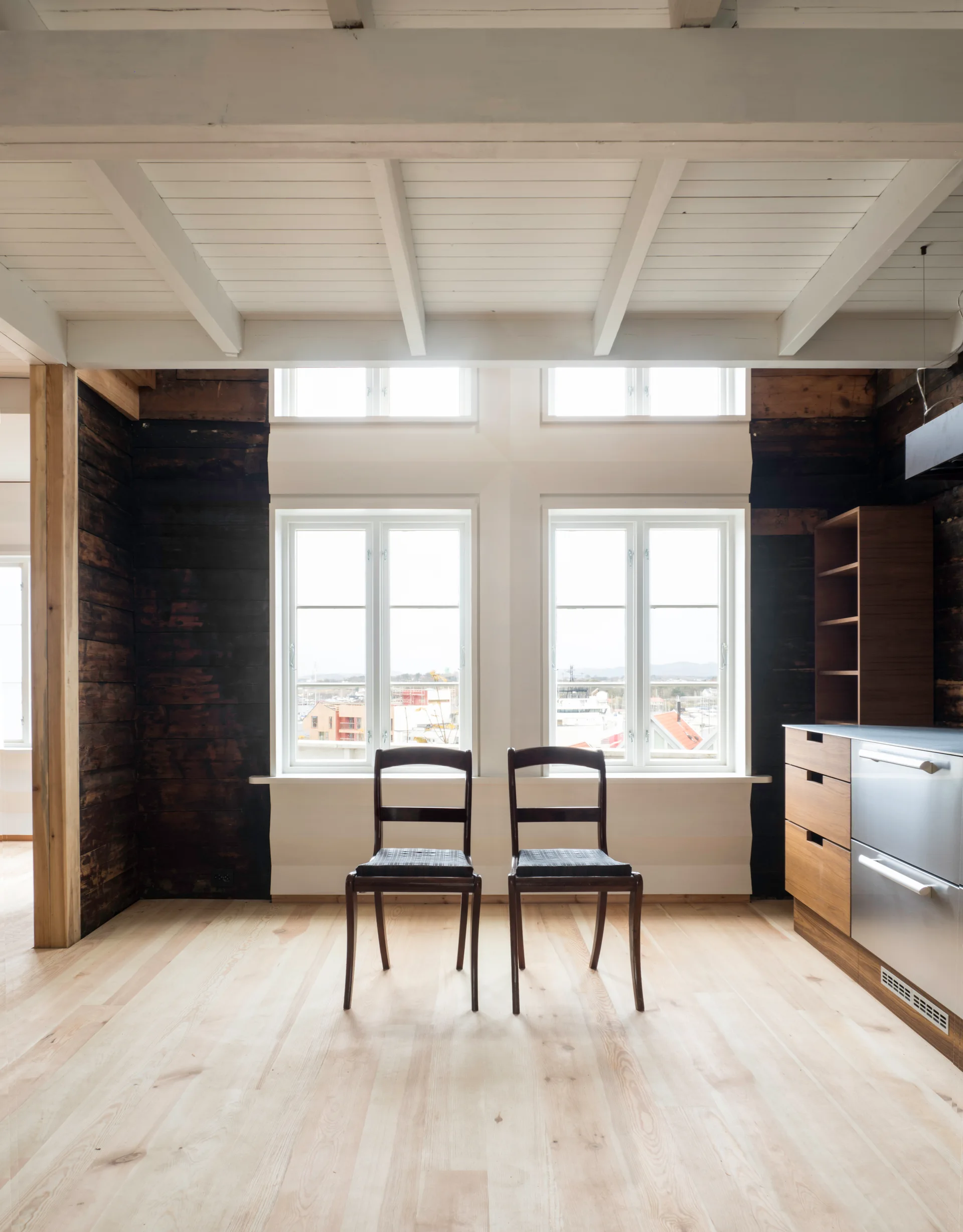 Two dark wood chairs stand centered in front of double windows in a room with dark wood walls and a white beamed ceiling.