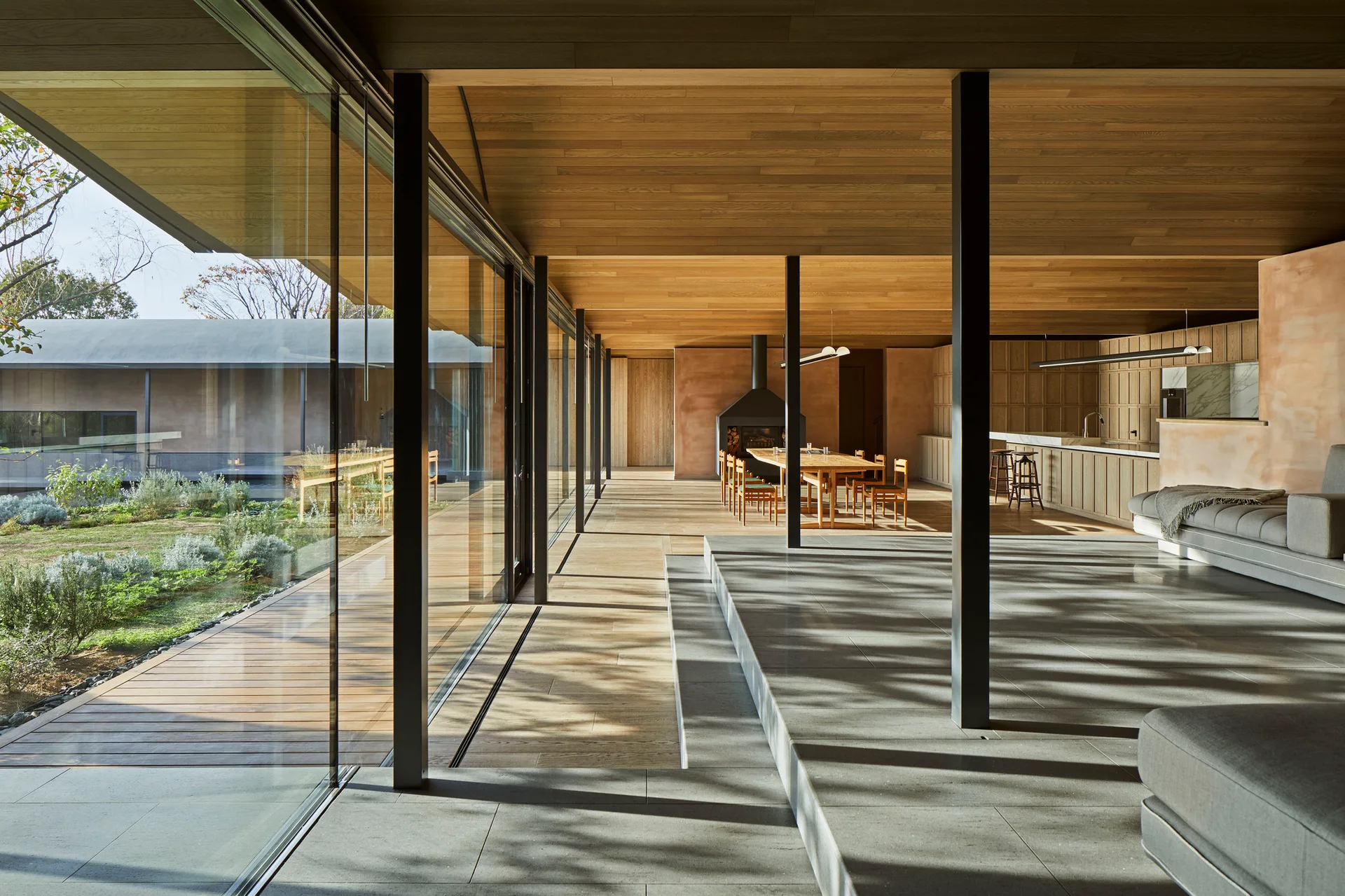 A minimalist open-plan living and dining space featuring a sunken stone floor, continuous wood ceilings, and large glass walls opening to a garden.