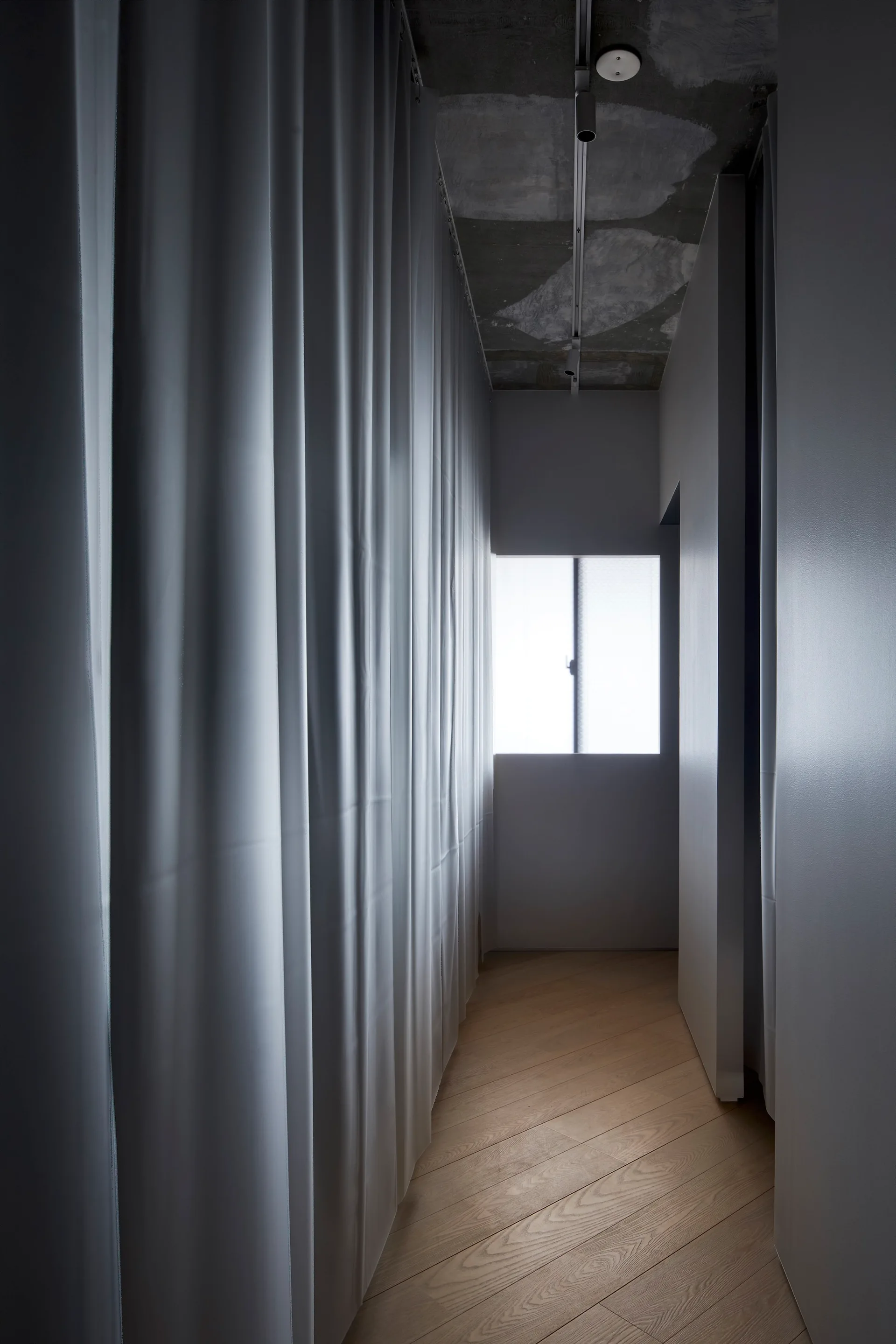 A narrow hallway featuring a wall of draped white curtains, diagonal light wood flooring, and an exposed concrete ceiling with track lighting.