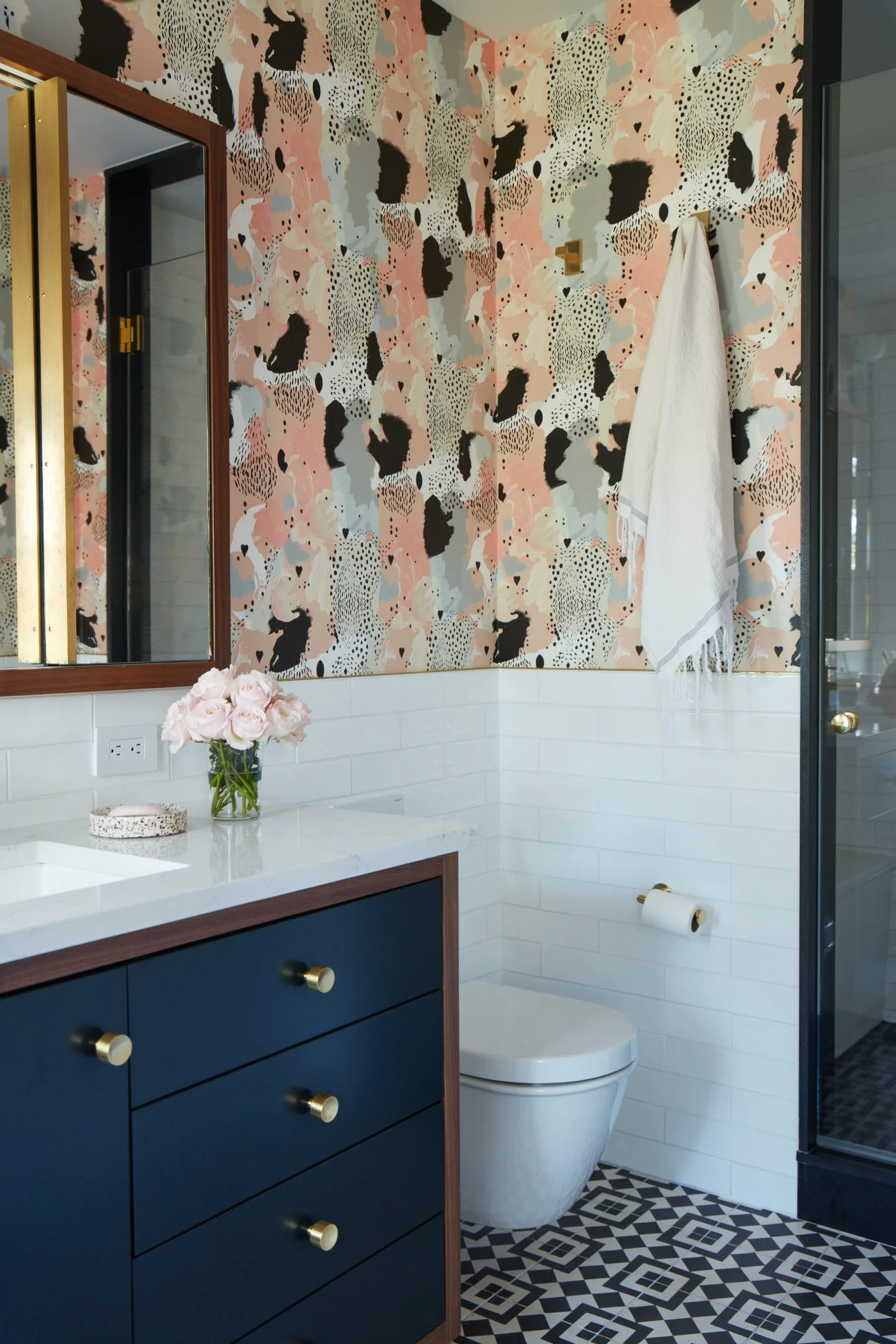 A bathroom with abstract pink and black wallpaper, white subway wall tile, a navy vanity with brass hardware, and a black and white geometric tile floor.
