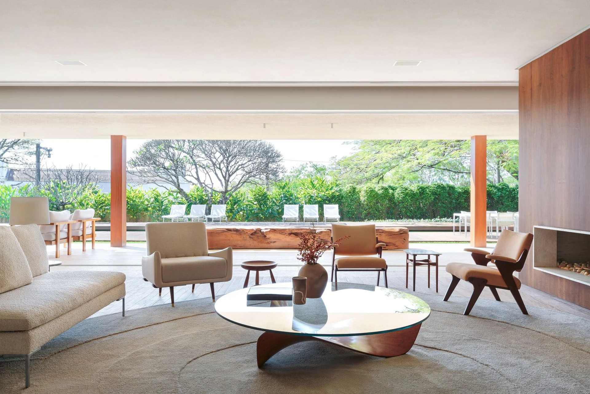 A mid-century modern living room with wood armchairs, a round glass coffee table on a circular rug, and a wide opening to an outdoor patio.