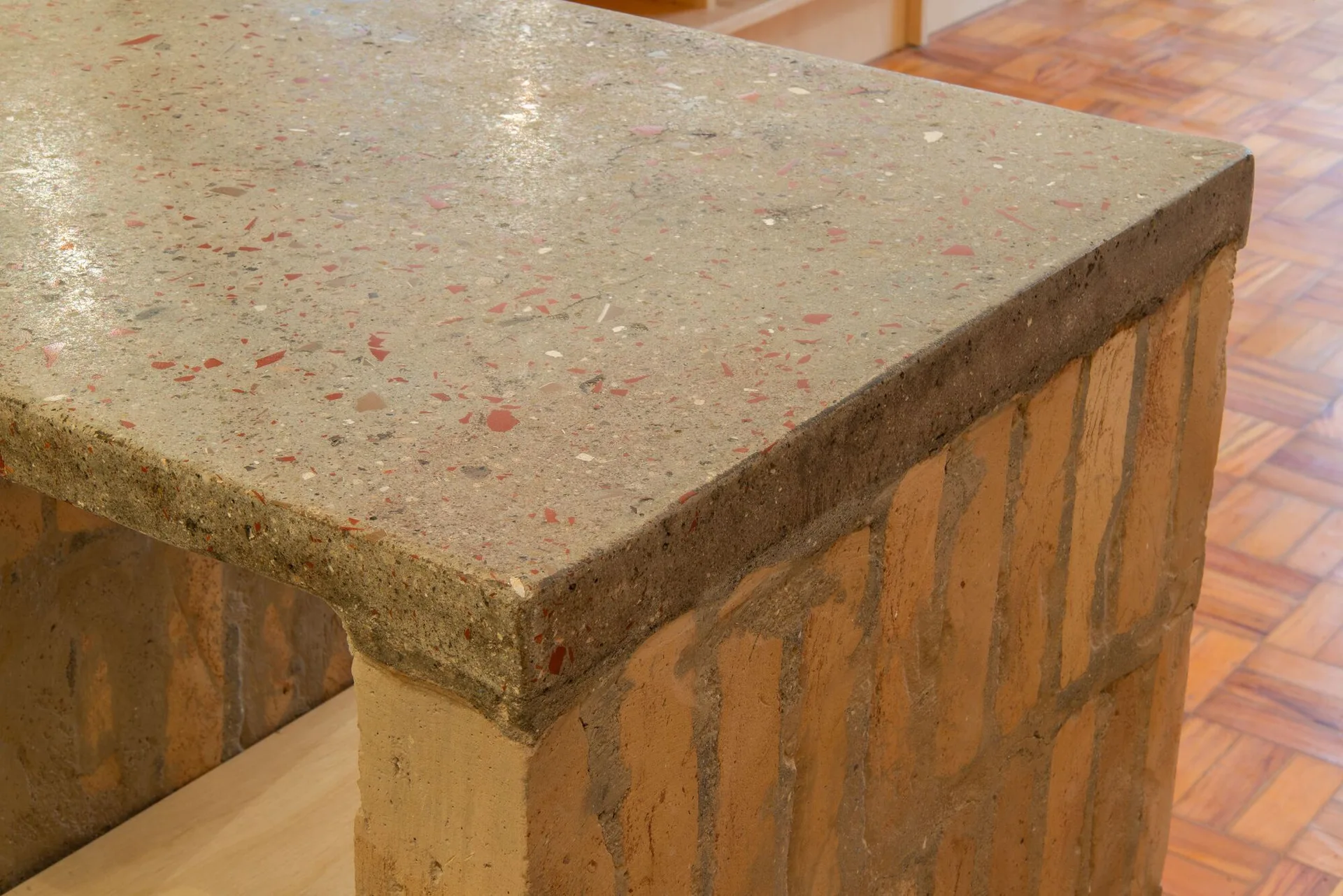 A close-up of a kitchen island featuring a speckled grey terrazzo countertop resting on a textured raw brick base over parquet floors.