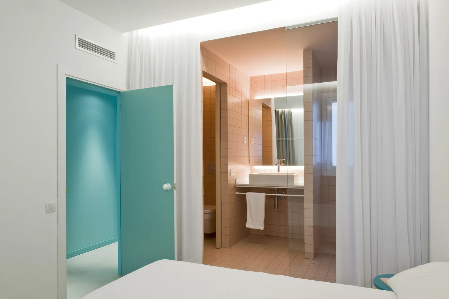 A bedroom view looking into a minimalist bathroom with pastel pink grid tiles, a floating vanity, sheer curtains, and a vibrant teal door.