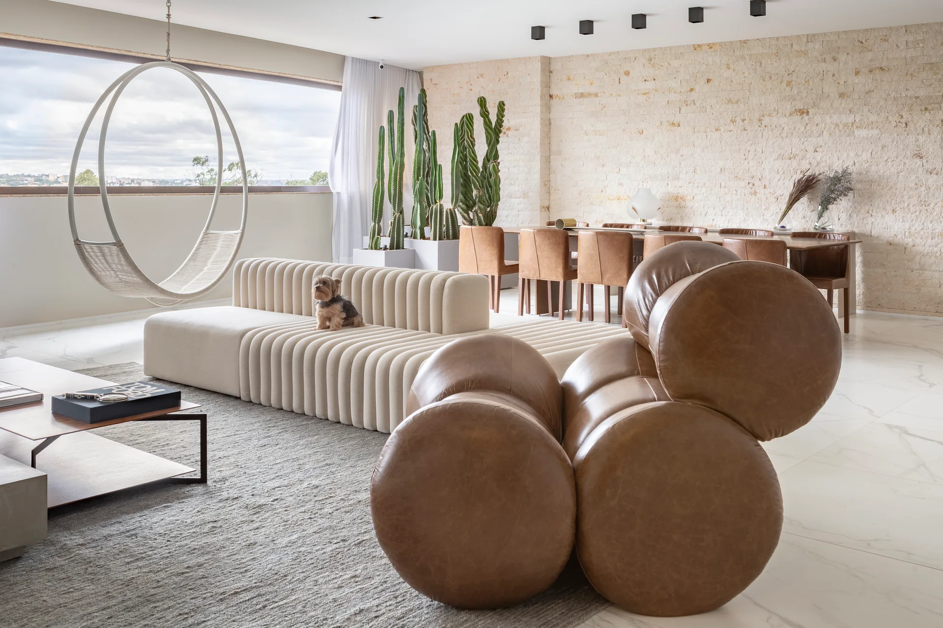 A contemporary living room features a ribbed cream sofa, a brown sculptural leather lounge chair, and tall cacti in front of a stone dining wall.
