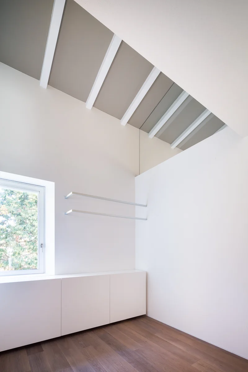 A minimalist white room features a sloped beamed ceiling, low-profile white cabinets, thin floating shelves, and a window overlooking green trees.
