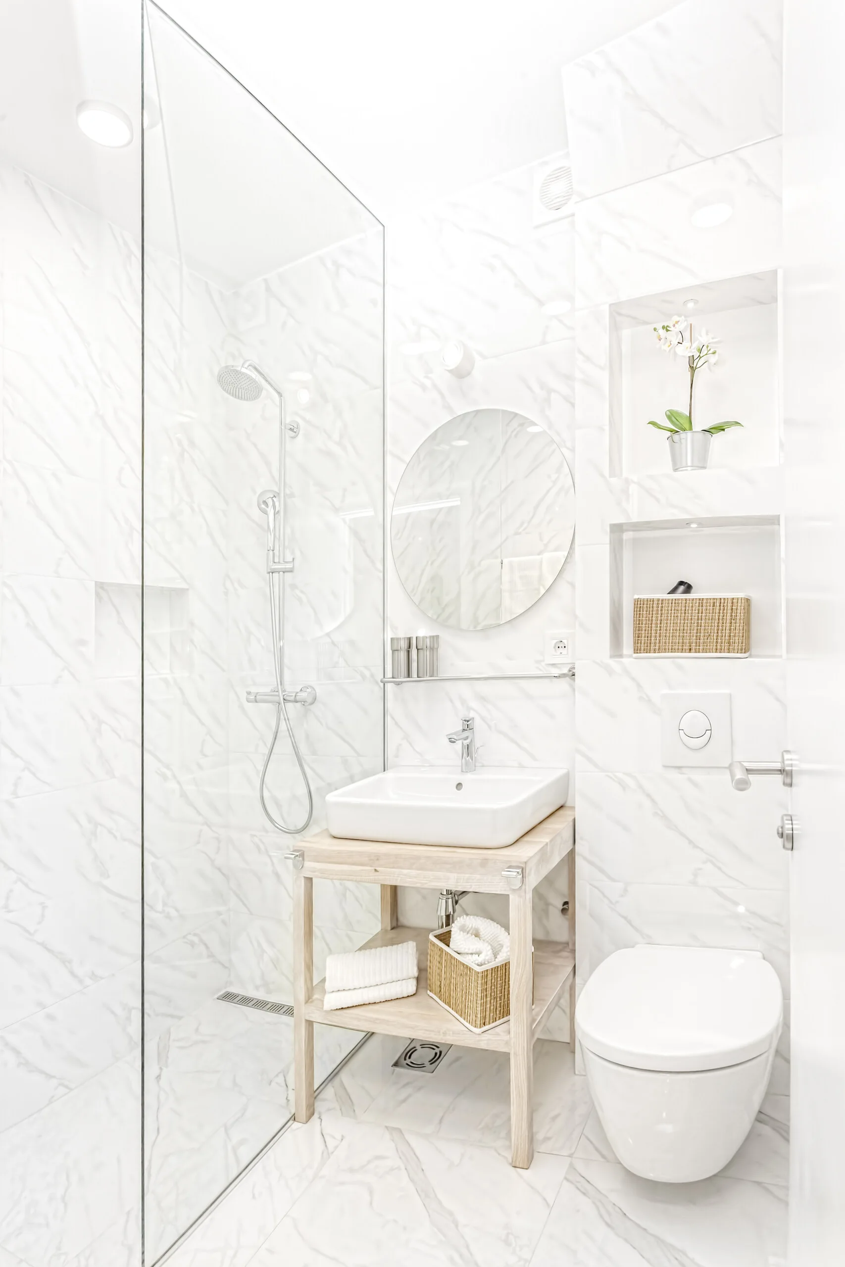 A modern bathroom features marble-patterned tiled walls, a light wood open vanity with a white vessel sink, a round mirror, and a glass shower panel.