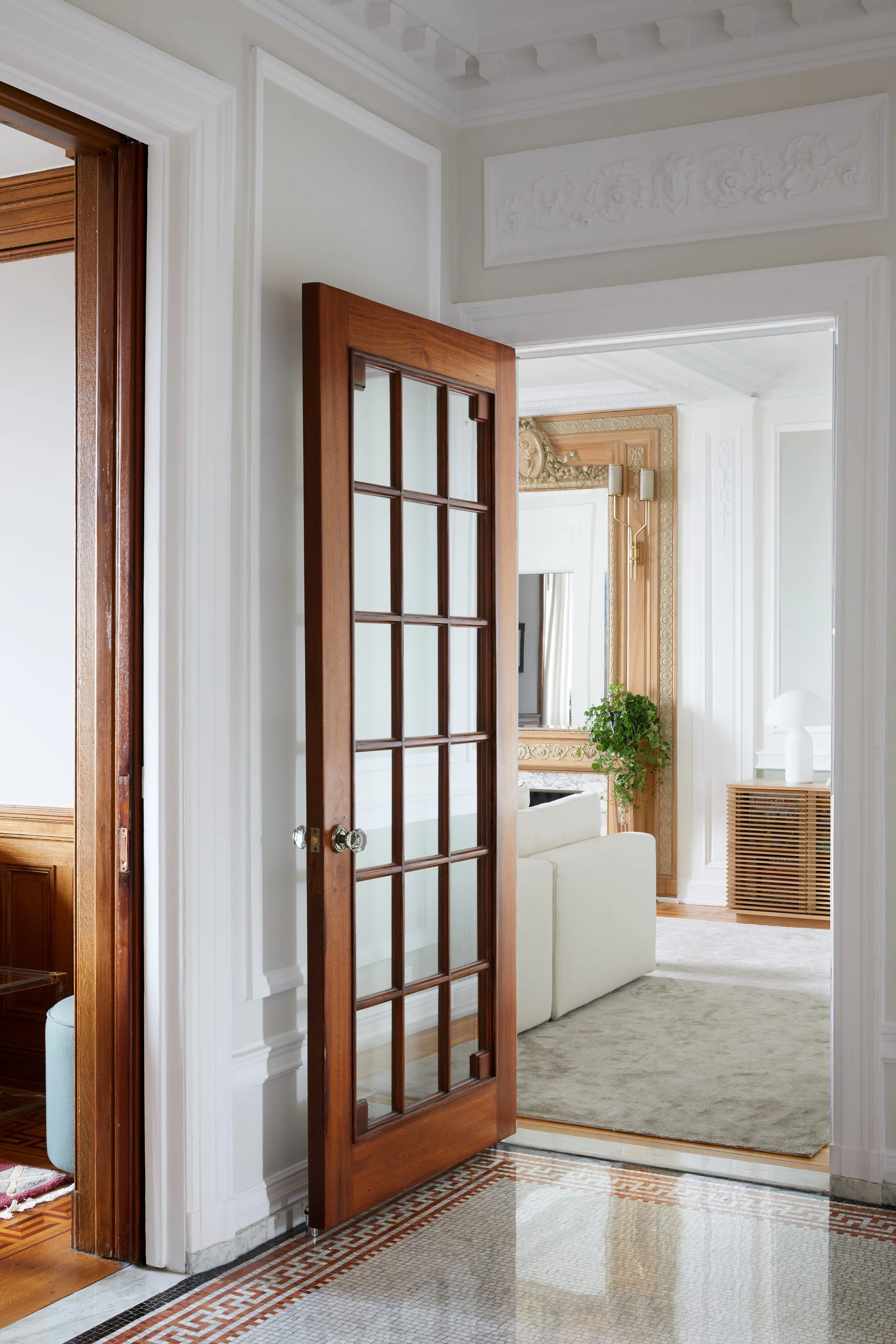 An open wood-framed French door leads from a mosaic tile hallway into a bright white living room with ornate wall moldings and a neutral sofa.