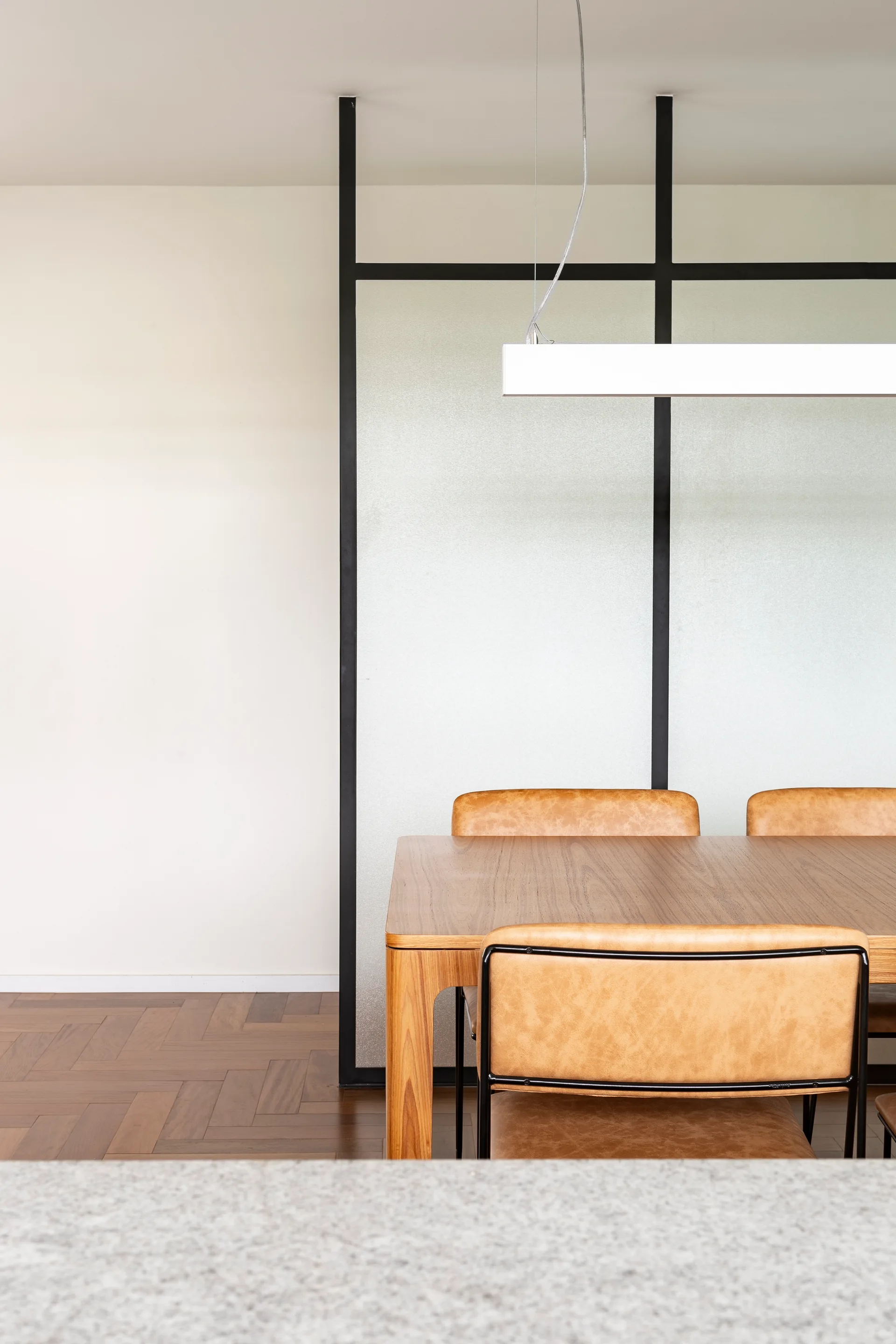 A wooden dining table paired with tan leather chairs sits in front of a black-framed frosted glass room divider on a herringbone wood floor.