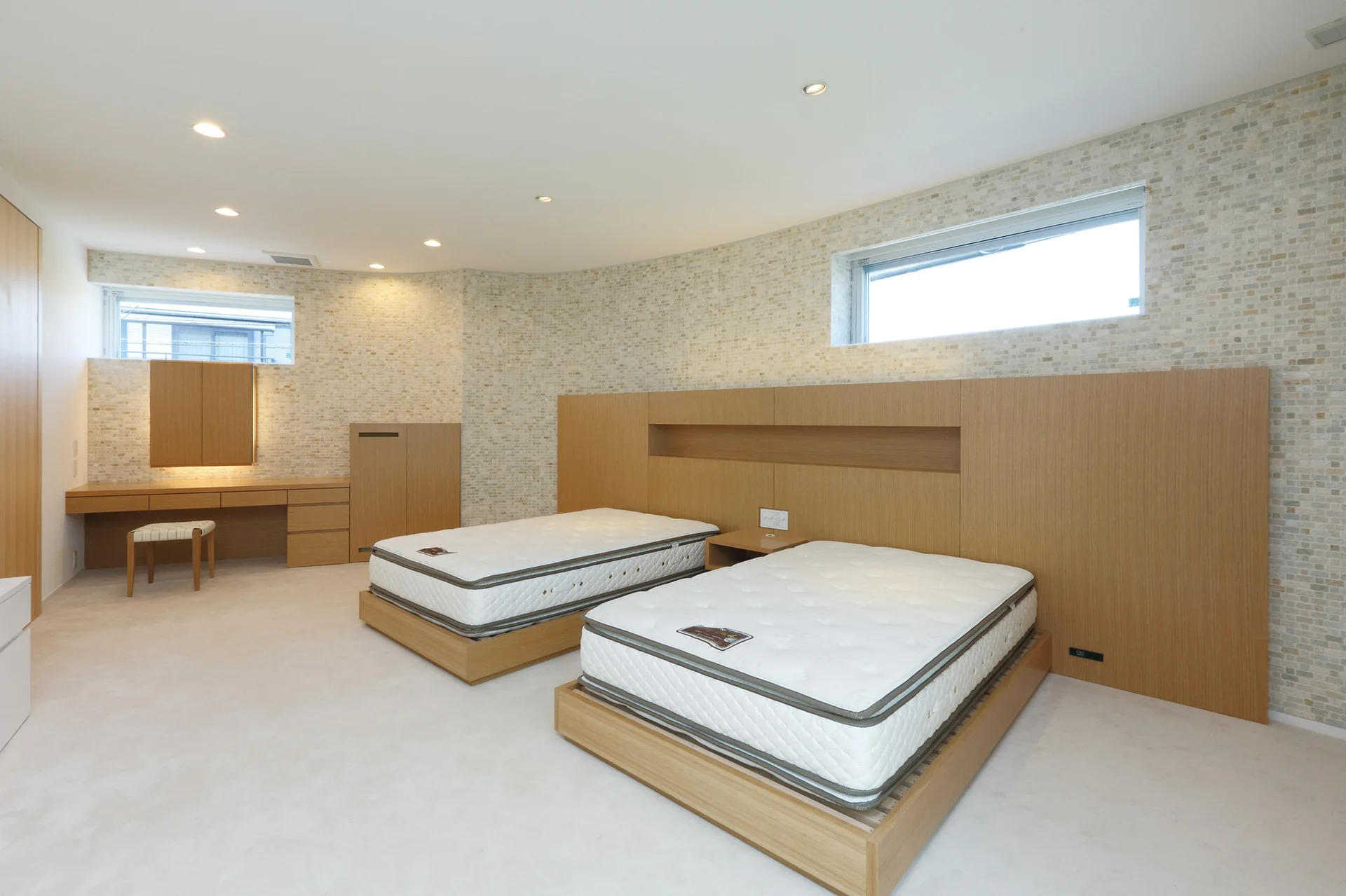 A minimalist bedroom featuring two beds, a custom wood headboard, and a built-in vanity desk against a neutral mosaic tile wall.