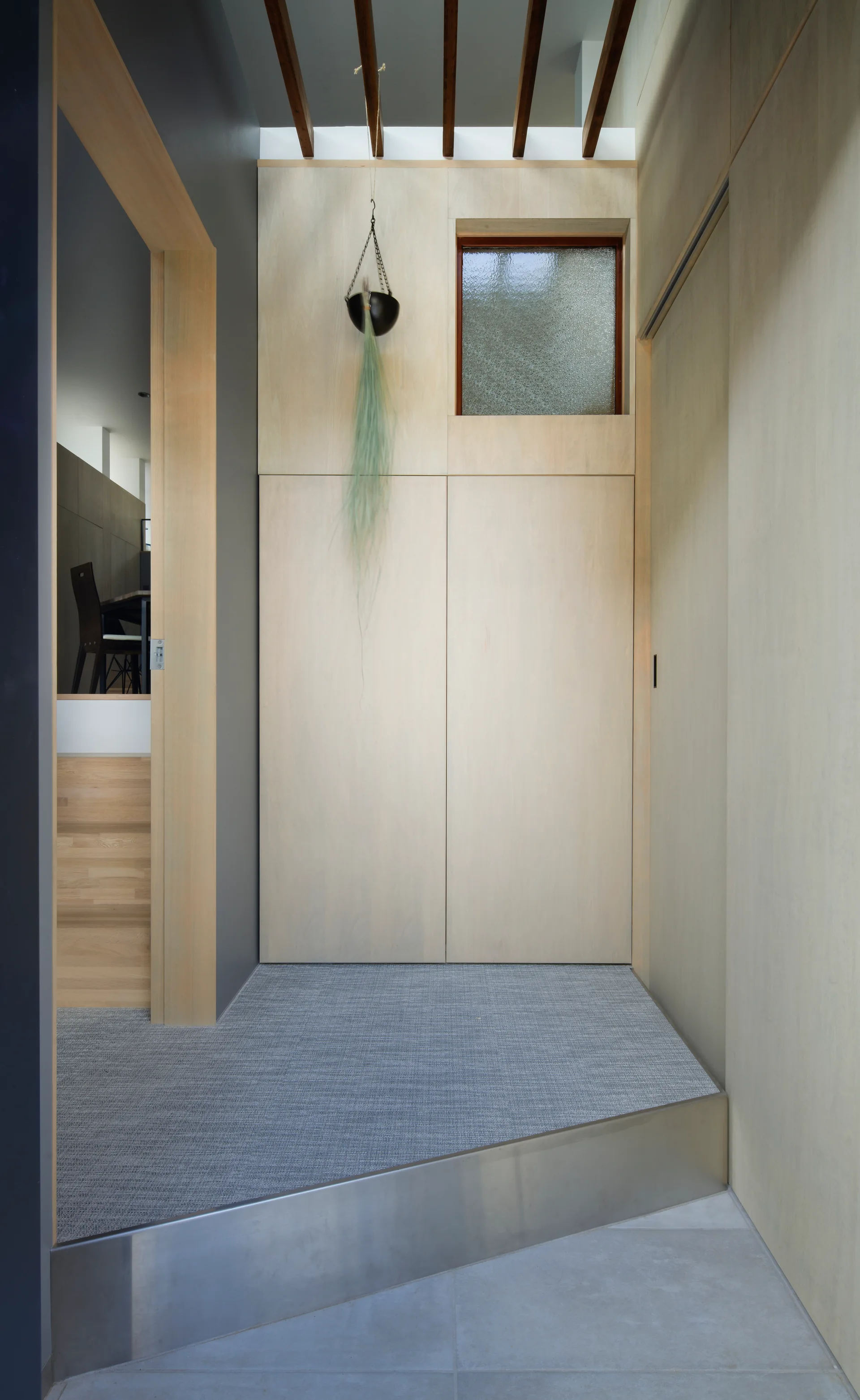 A modern minimalist entryway features light plywood wall paneling, a gray stepped floor with a metal threshold, and dark ceiling beams.