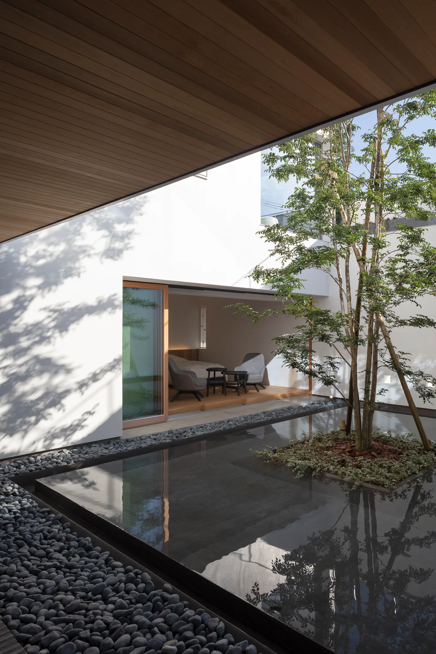 A modern minimalist courtyard featuring a shallow dark pool with a single tree surrounded by pebbles and white exterior walls.