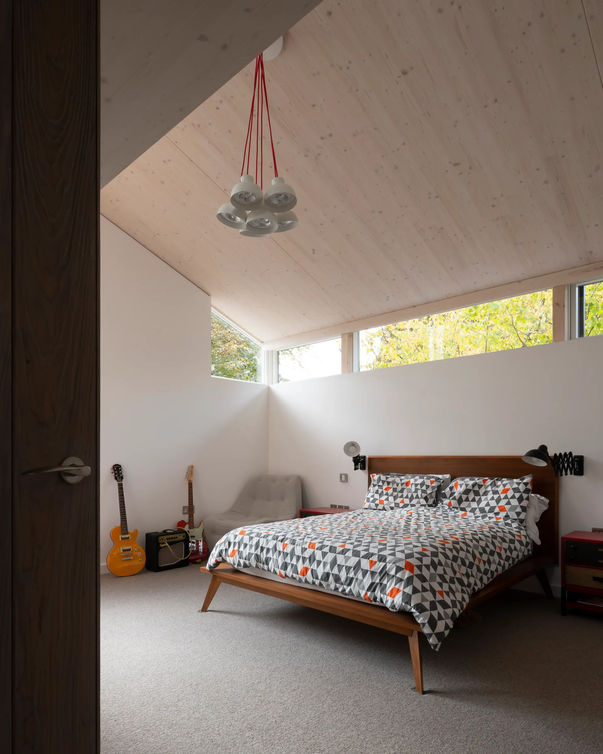 A modern bedroom features a wooden bed with geometric bedding beneath a sloped timber ceiling, accented by horizontal clerestory windows.