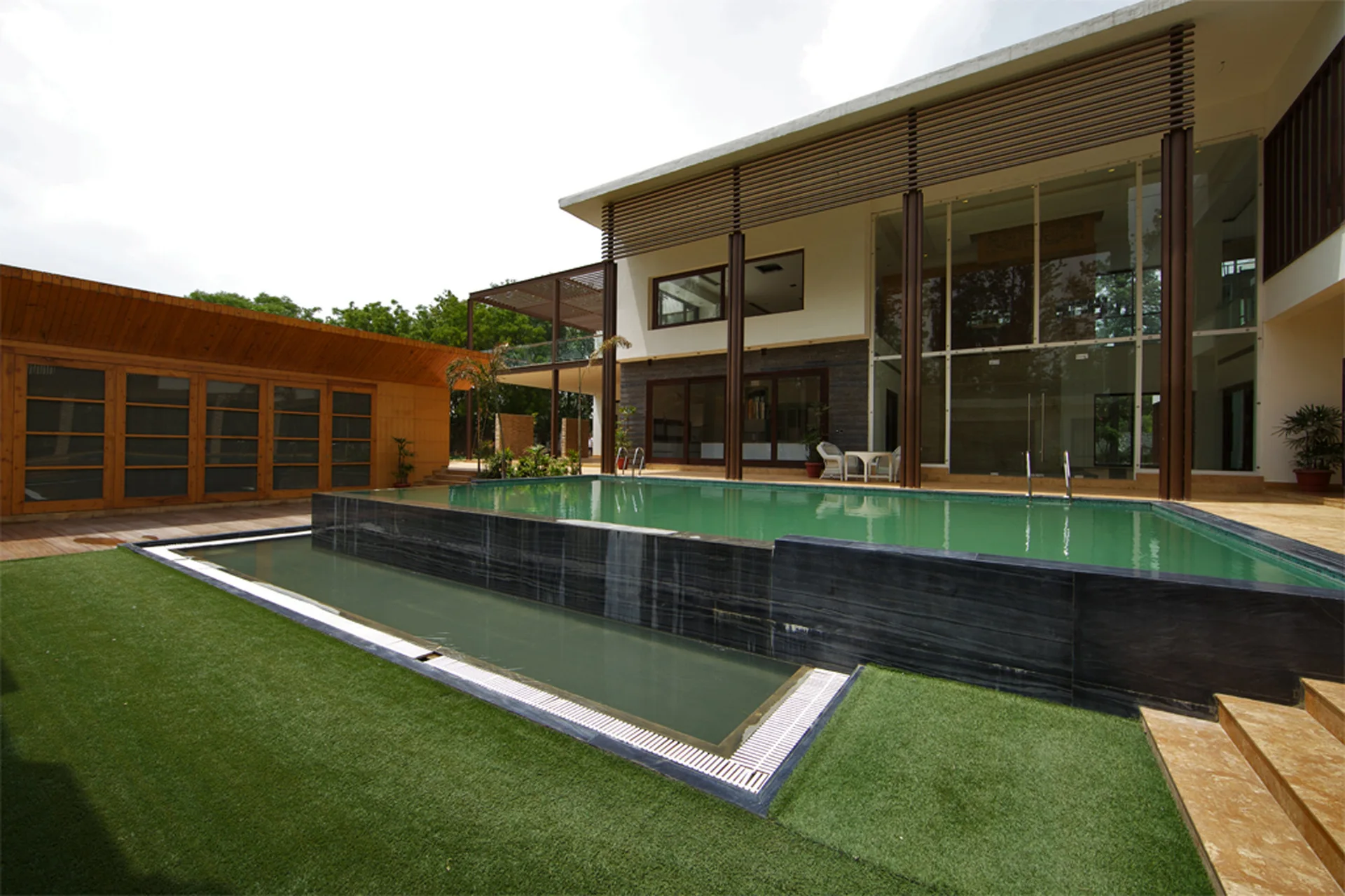 A two-story modern house with a large rectangular swimming pool bordered by dark stone and a green lawn under a bright sky.