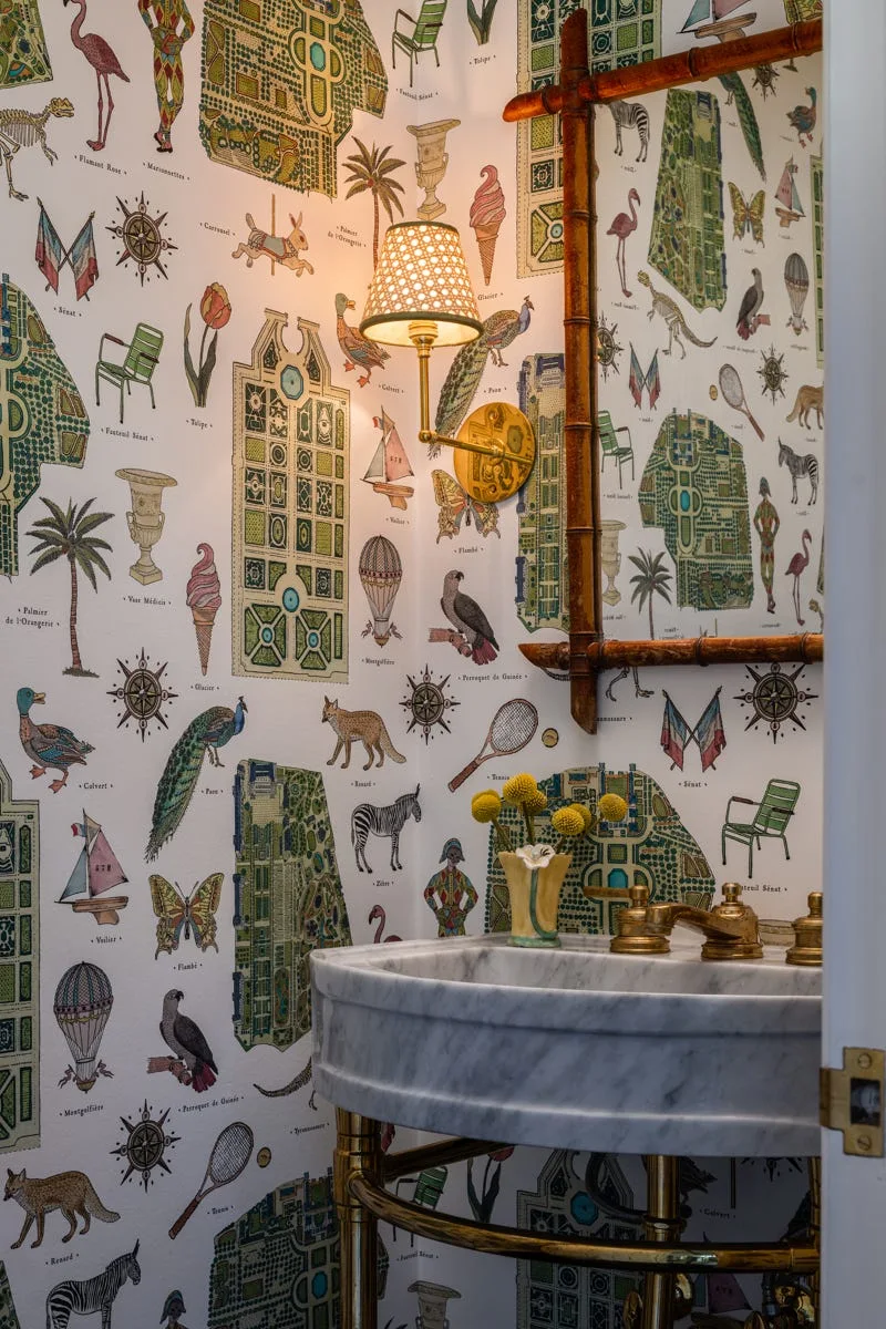 A powder room features detailed illustrated wallpaper behind a marble sink with gold fixtures, a warm brass sconce, and a bamboo-style mirror.