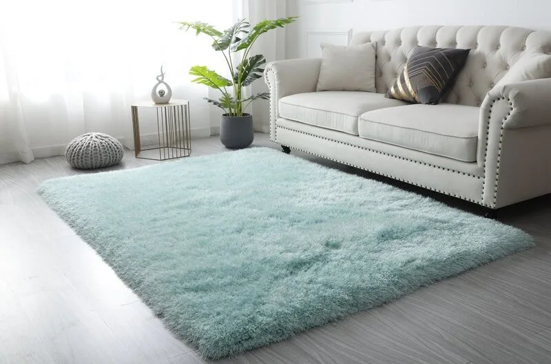 A light blue shaggy area rug anchors a living room featuring a tufted off-white sofa, a gold side table, and a potted houseplant near a bright window.