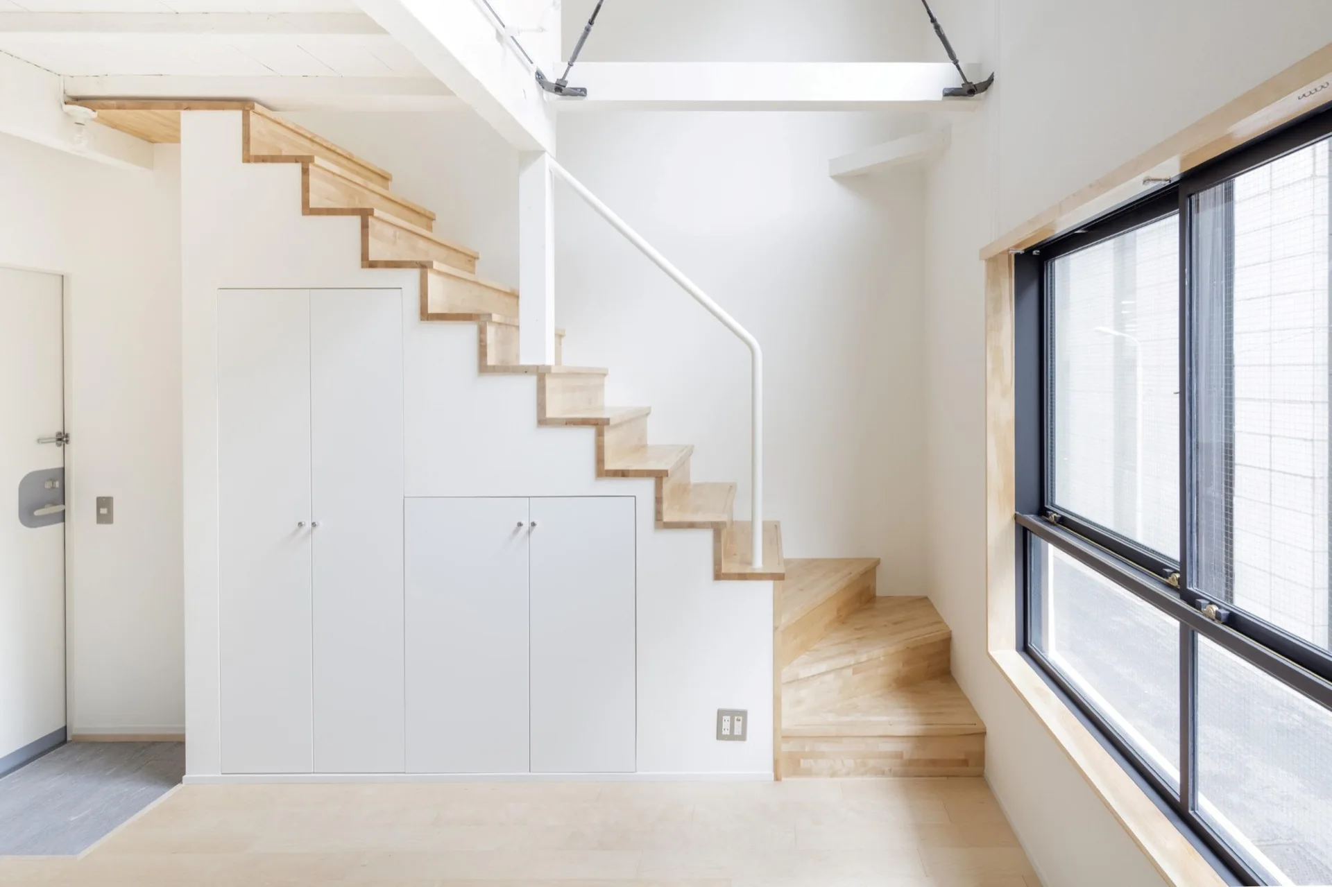 A minimalist staircase with light wood treads features white built-in storage cabinets underneath and a black-framed window on the side.