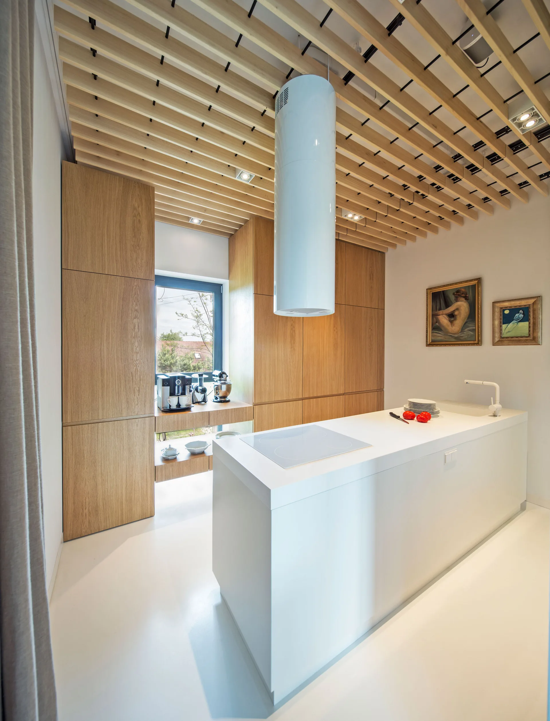 A modern kitchen features a central white island under a white cylindrical range hood, with wood cabinets and a slatted wood ceiling.
