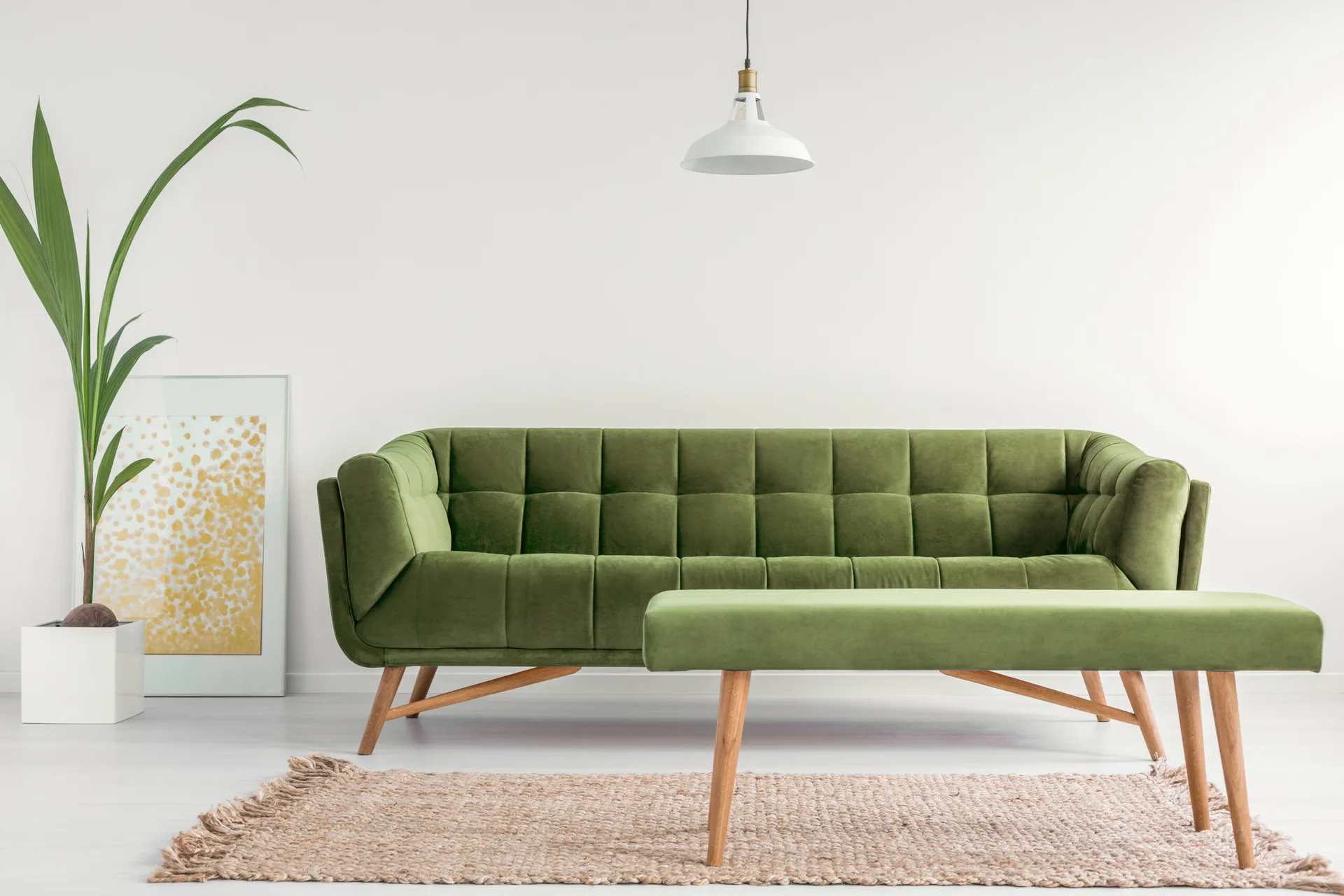 A minimalist living room featuring a green tufted velvet sofa, matching green bench, woven jute rug, and a tall plant against a white wall.