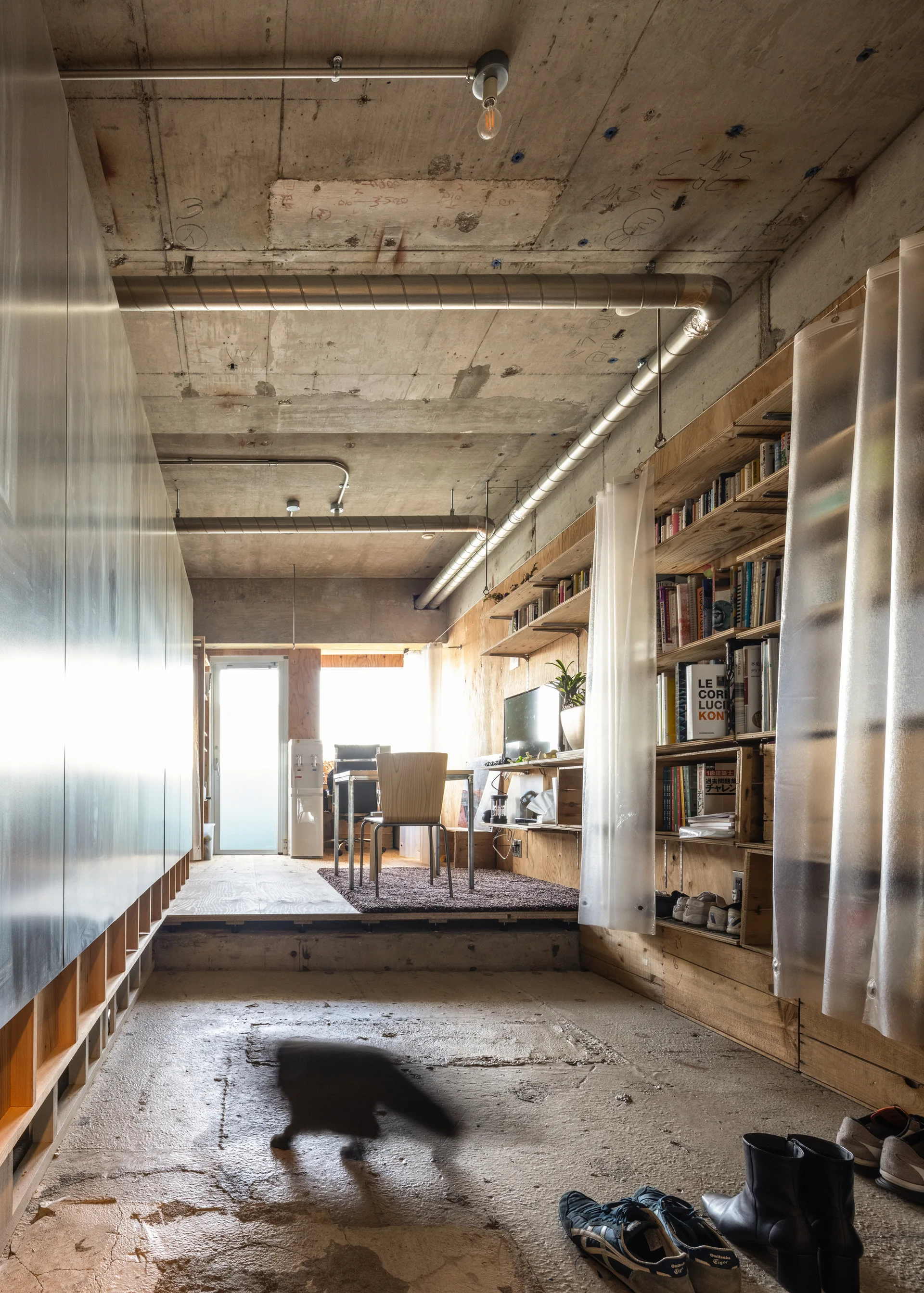 An industrial home office features an elevated plywood platform floor, unfinished concrete ceiling, and a large wooden shelving unit with translucent curtains.