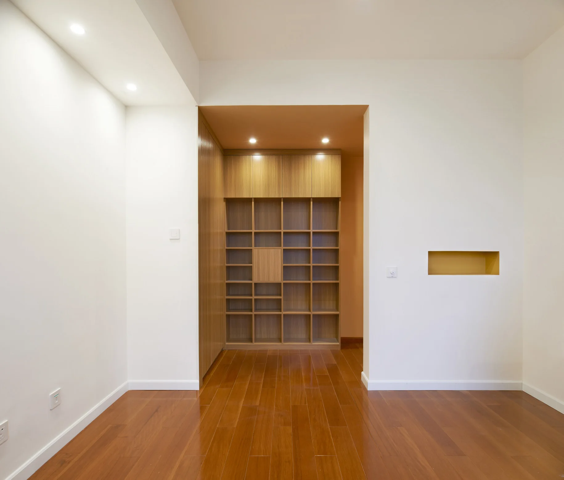 An empty room with polished hardwood floors features a custom wooden shelving unit built into a recessed wall alcove.
