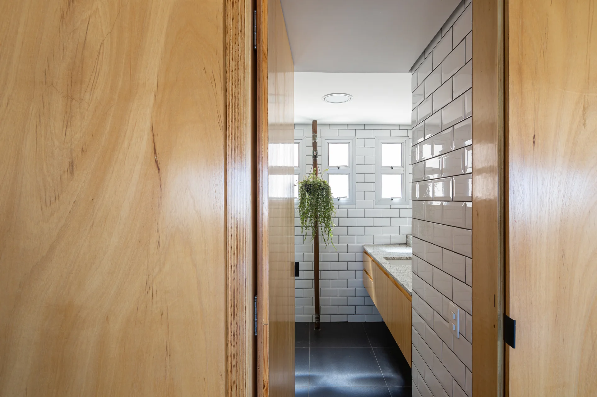 A view through a wooden doorway into a bathroom with white subway tile walls, a light wood floating vanity, and dark gray floor tiles.