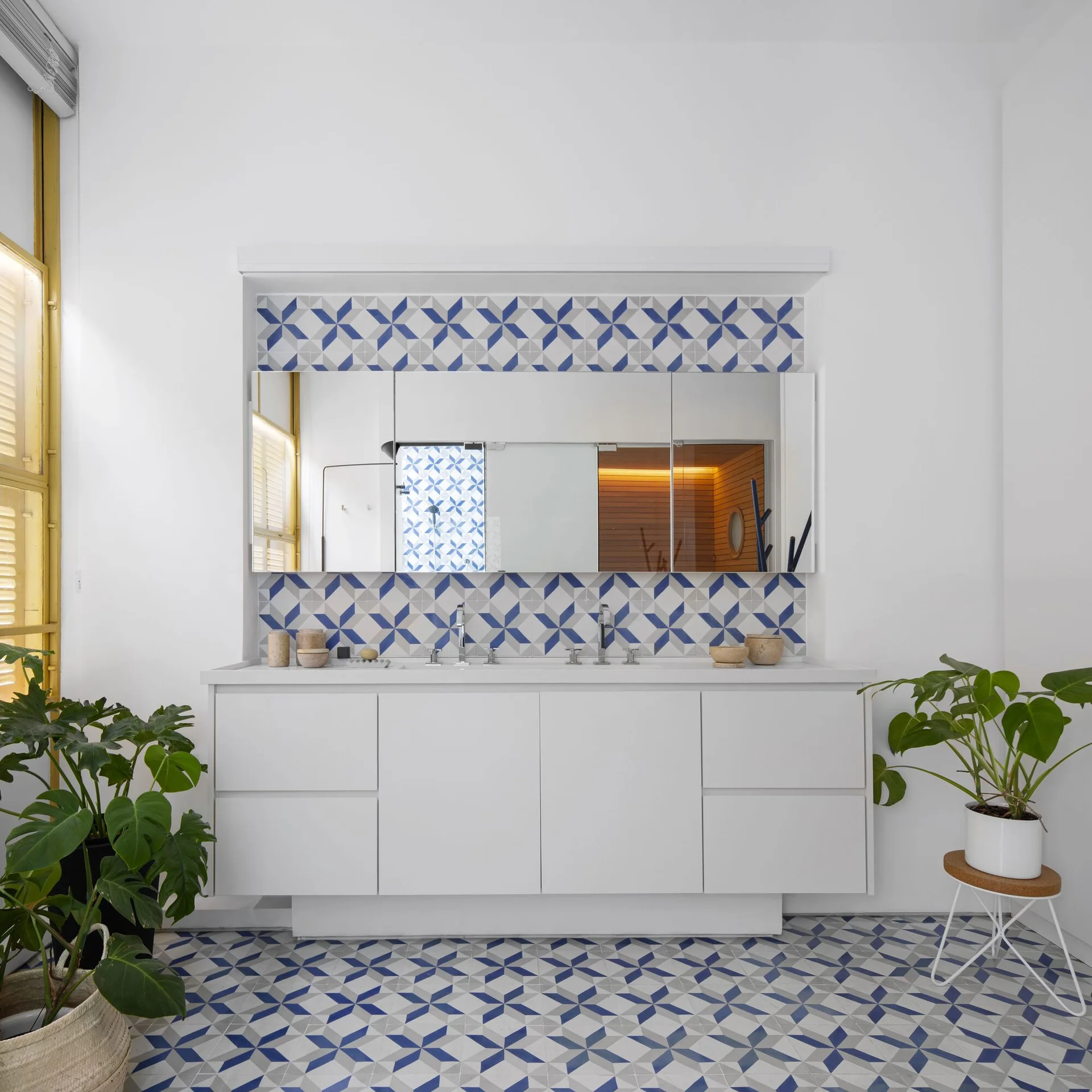 A white double vanity bathroom features matching blue and grey geometric tiled floors and backsplash, framed by green potted plants.