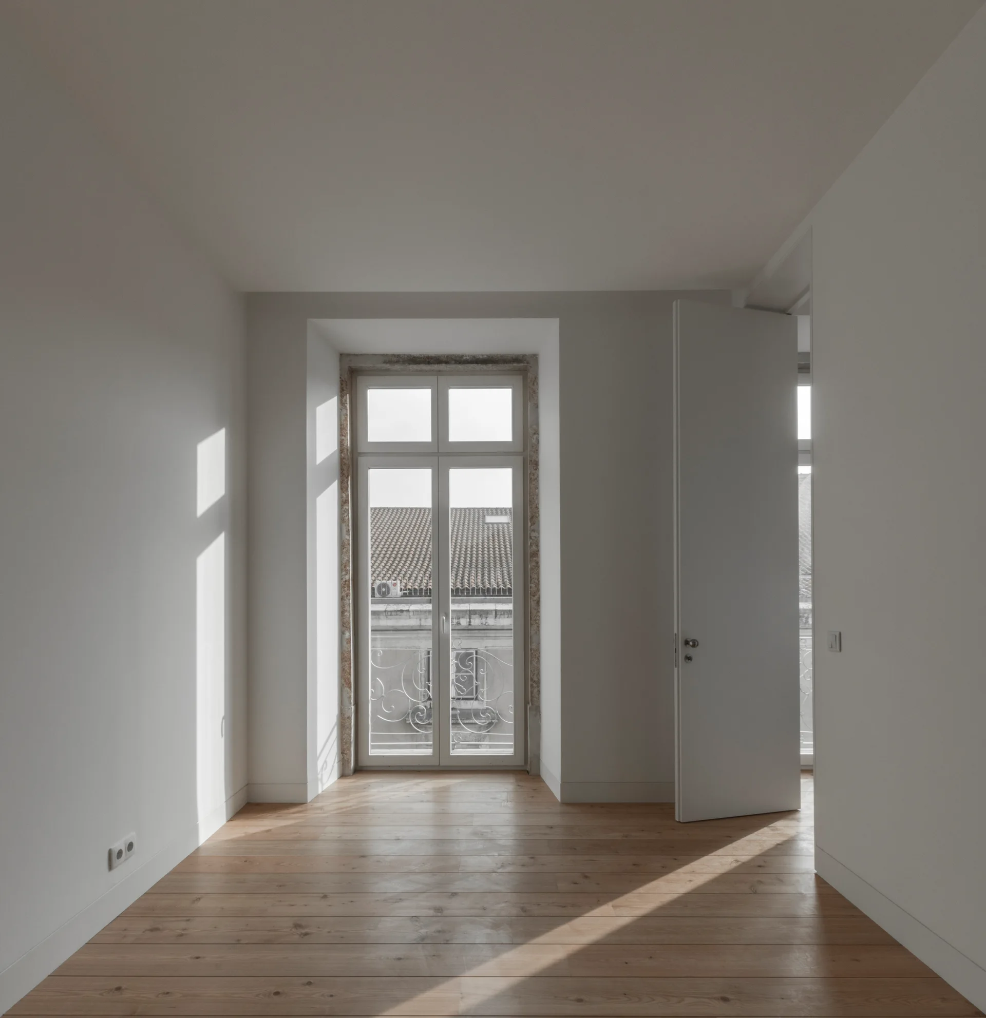 An empty room with white walls, wide plank hardwood floors, and a tall French window casting diagonal sunlight across the space.