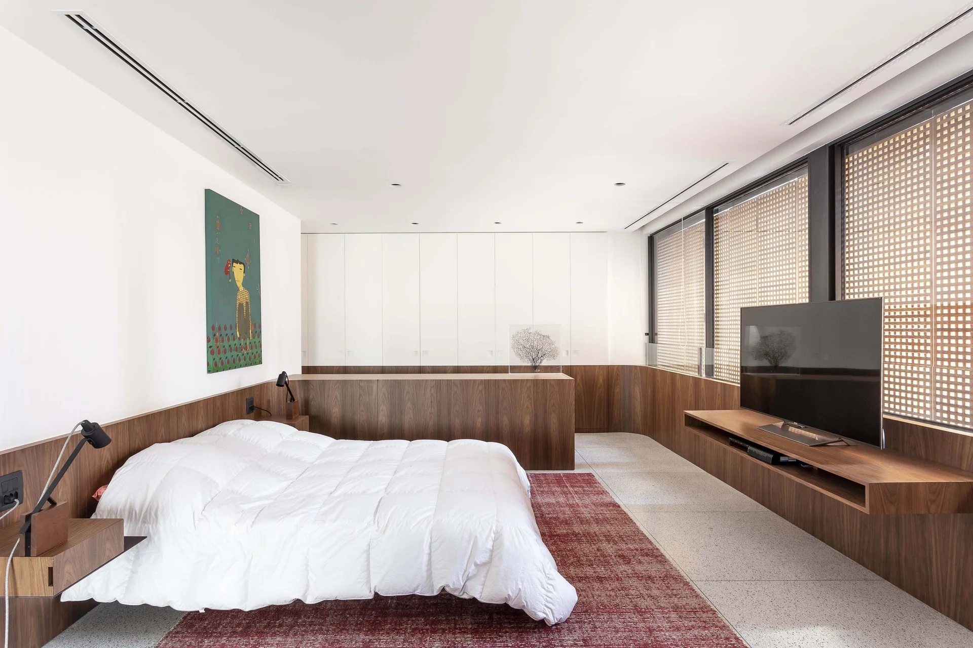 A modern bedroom features a white bed on a red rug, surrounded by warm wood wainscoting, a floating media console, and built-in white cabinets.