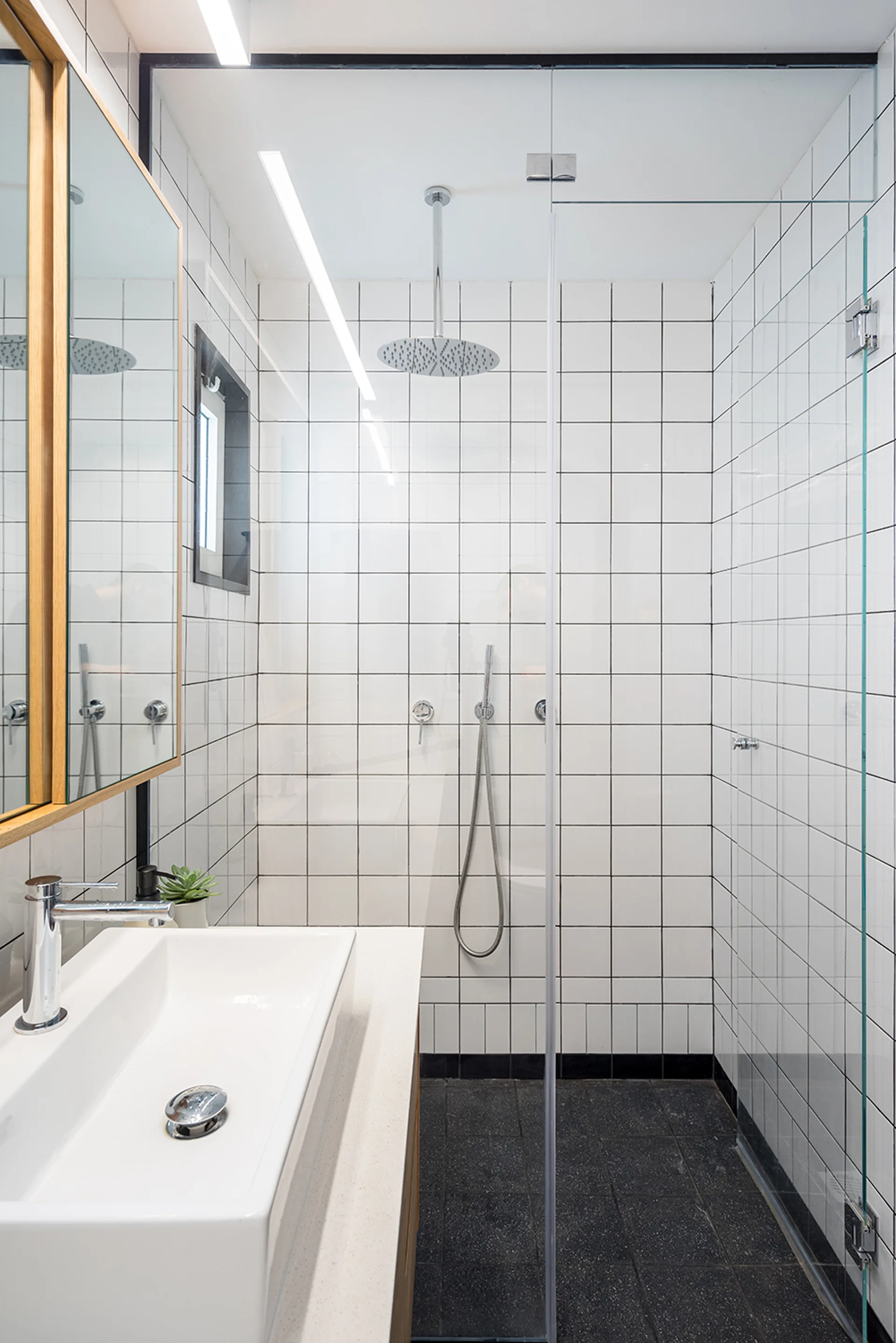 A modern bathroom features white square-tiled walls with dark grout, a walk-in glass shower, and a wood-framed vanity mirror.