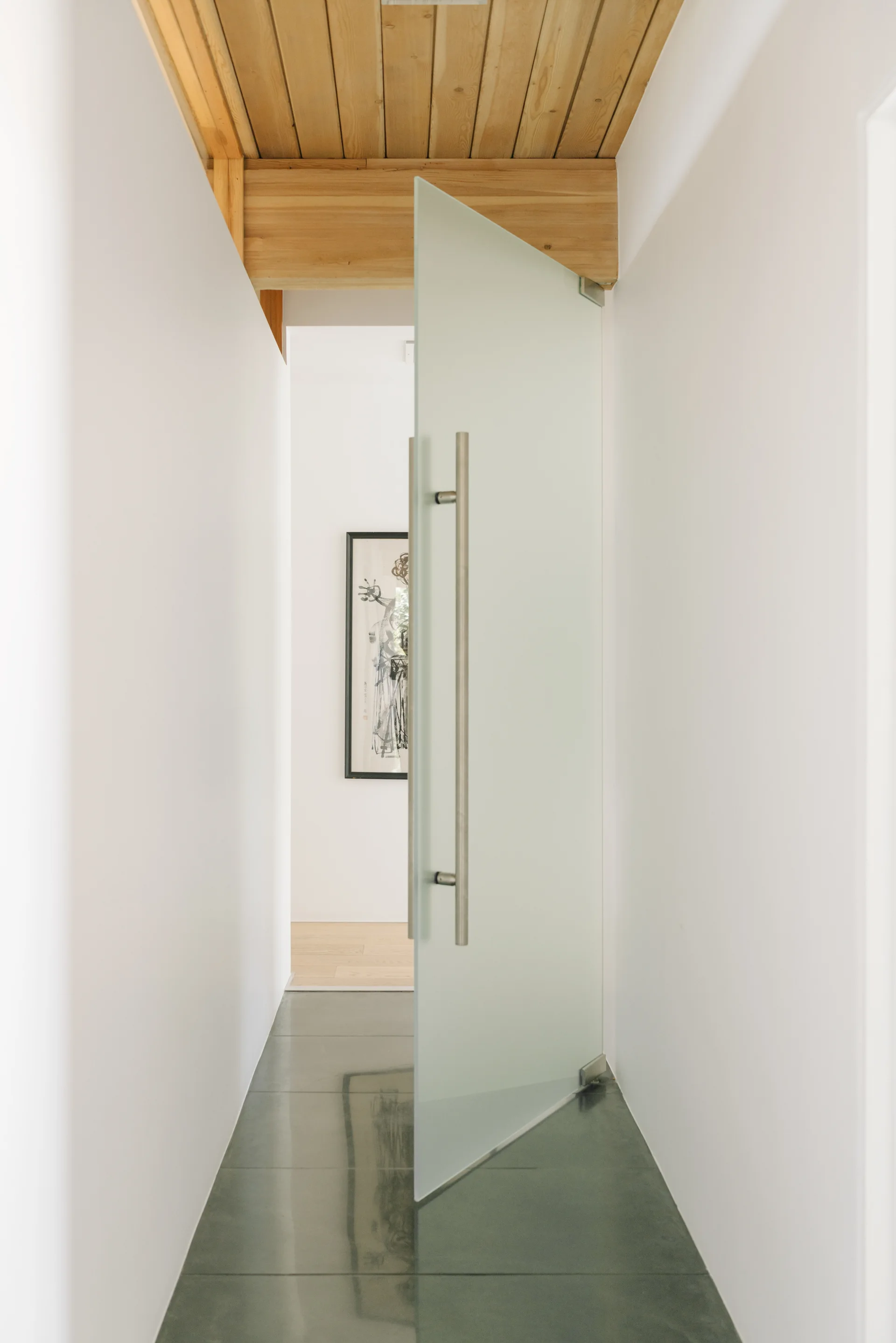 A modern hallway features a pivoting frosted glass door with a long metal handle, a natural wood plank ceiling, and grey tiled floors.