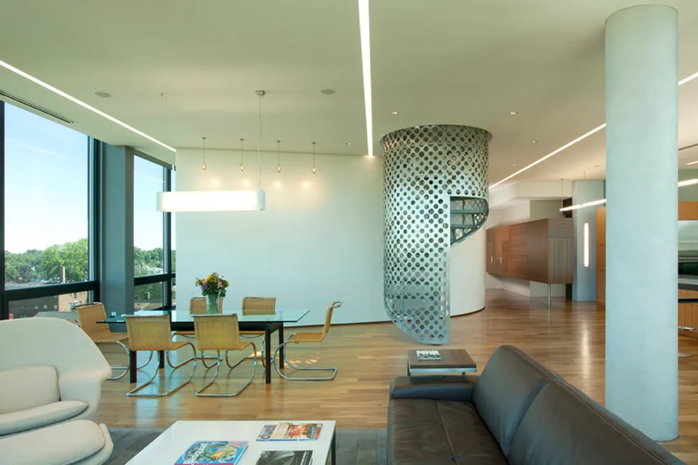 A modern open-plan living and dining space features a perforated metal spiral staircase, a glass table, a leather sofa, and wood flooring.