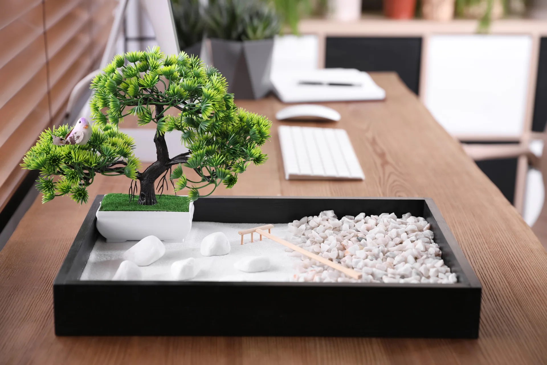 A miniature desktop zen garden with white sand, a small artificial bonsai tree, and tiny stones rests on a wooden home office desk.