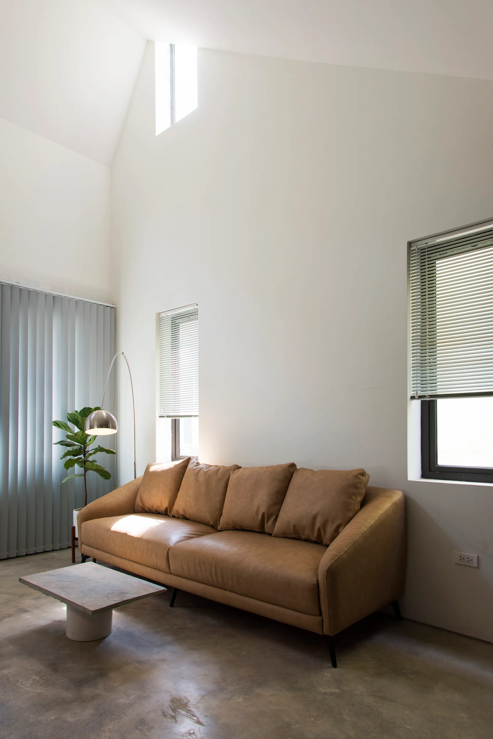 A tan leather sofa sits on a polished concrete floor beneath a tall white vaulted ceiling in a minimalist living room.