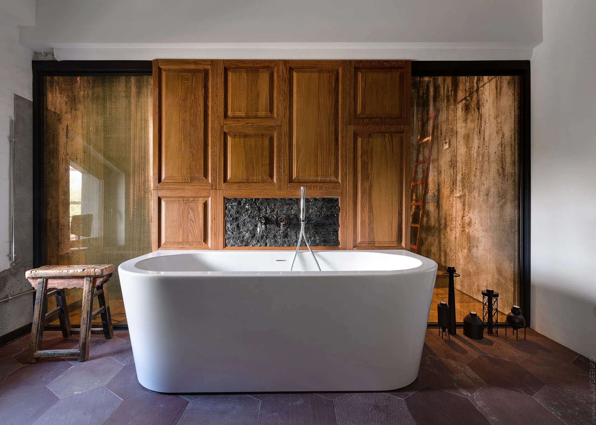 A white freestanding bathtub sits on dark hexagonal floor tiles in front of a warm wood-paneled wall with a textured stone insert.