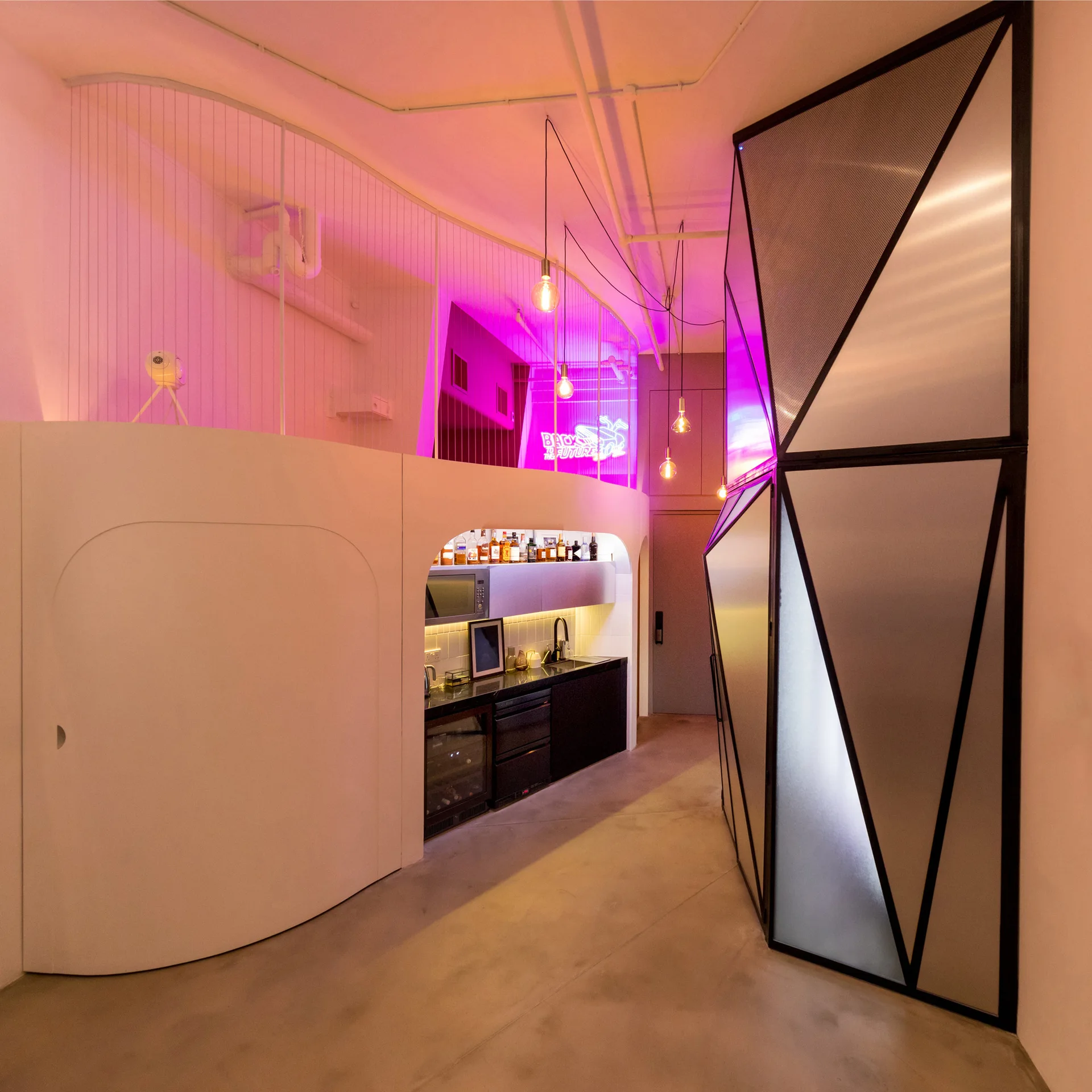 A modern home bar tucked under a white curved loft features a sink, wine cooler, hanging pendant lights, and a faceted black geometric screen.
