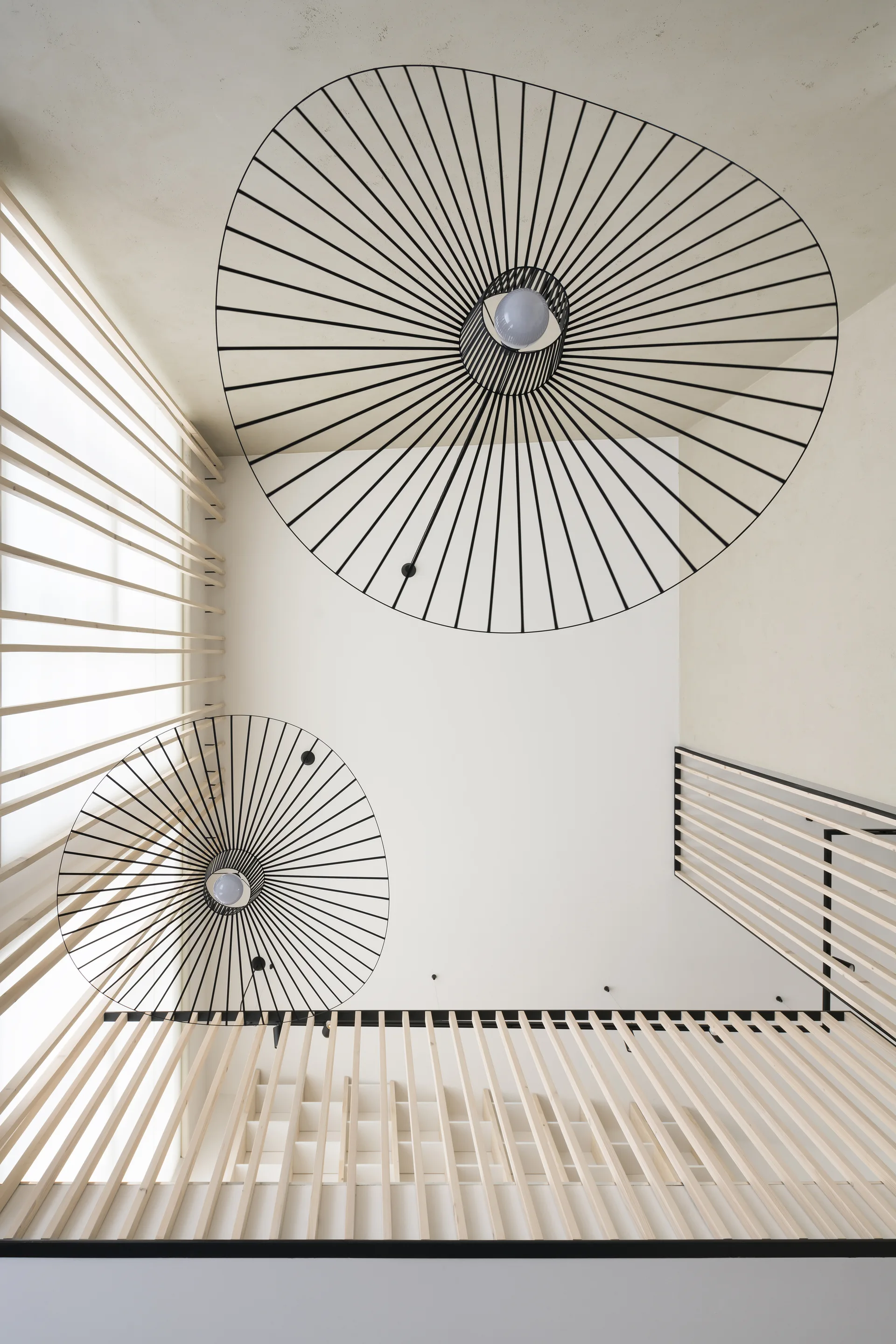 Looking up a modern stairwell featuring vertical light wood slats and two large black circular wire pendant lights hanging from a white ceiling.