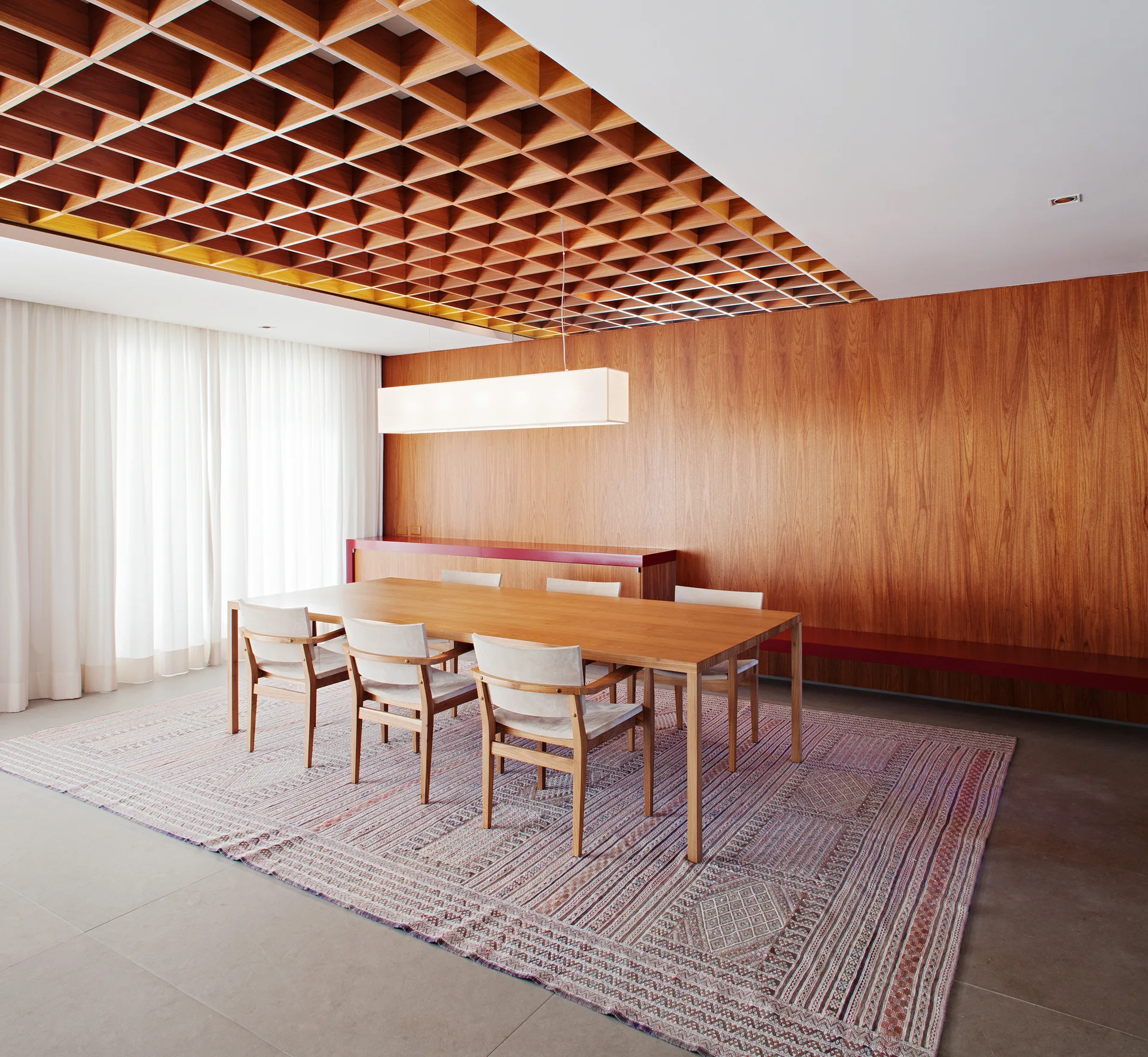 A modern dining room features a long wooden table, light chairs, a patterned rug, a wood-paneled wall, and a dramatic grid ceiling.